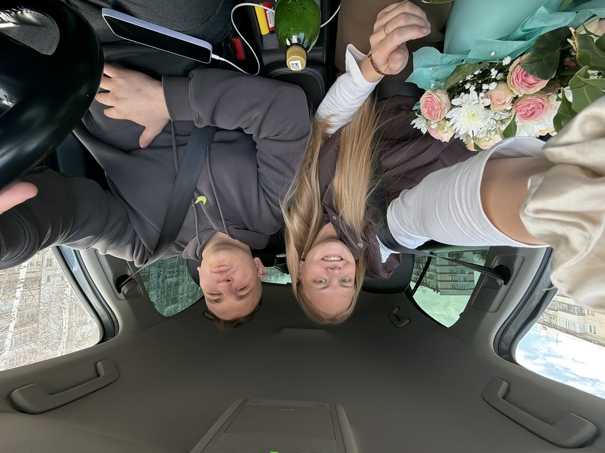 The image is a selfie taken from a low angle inside a car, looking upwards at two people in the front seats. The photograph is oriented vertically, causing the subjects to appear upside down from a conventional viewing perspective.
At the top center of the frame, the faces of a man and a woman are visible, both smiling. The man, positioned on the viewer's left (driver's side), has short dark hair and a slight wink or squint. He wears a dark gray hoodie with a small light green Nike swoosh logo visible on his left chest. A black seatbelt crosses his body. His left hand is seen near the steering wheel.
The woman, on the viewer's right (passenger's side), has long blonde hair flowing downwards towards the center of the image. She wears a brown jacket over a white long-sleeved top. A gold ring is on her left ring finger and a brown bracelet on her right wrist. She holds a bouquet of flowers, which includes pink roses, white chrysanthemums, and baby's breath, wrapped in light blue tissue paper.
Between the two individuals, towards the bottom of their torsos in the image, a white smartphone in a light blue case is charging via a white cable, resting on the car's console or dashboard. Next to it is a red object (possibly a power bank) and a green glass bottle with a gold cap, which has a small white sticker featuring an illegible black code.
The car's gray interior, including the headliner, parts of the dashboard, and sections of the windows, frames the subjects. Through the windows, bare trees and multi-story buildings can be seen, indicating an urban or suburban setting under daylight. The expressions of the man and woman convey happiness, suggesting a pleasant moment or celebration, possibly involving a gift.