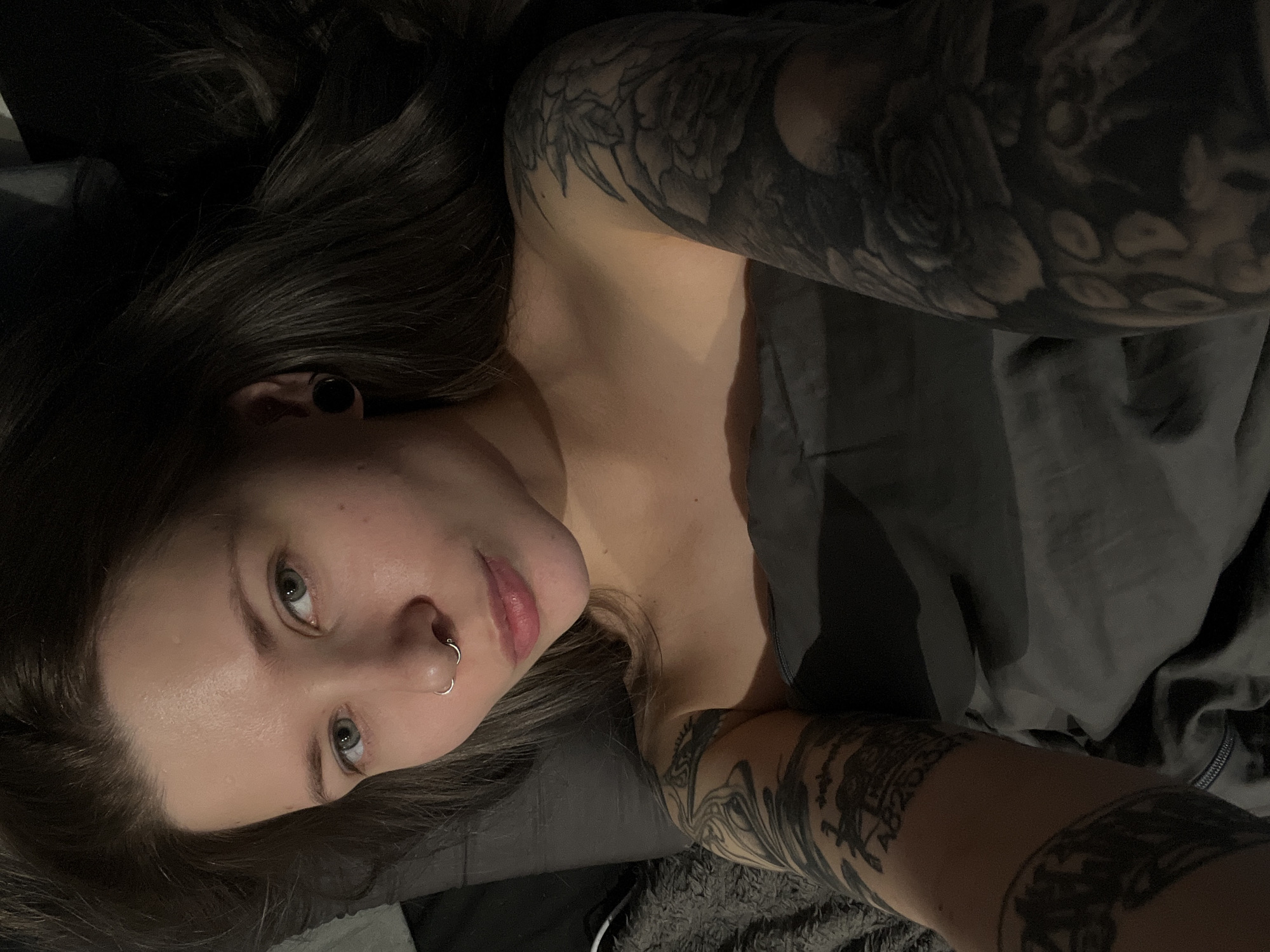 A woman is lying on a bed. She has fair skin, brown hair, green eyes, and a nose ring. Her arms are heavily tattooed in black and grey. She is covered with a grey blanket. The image appears to have been taken indoors in dim lighting. There are no discernible indicators of location within the city of Khimki, Russia.