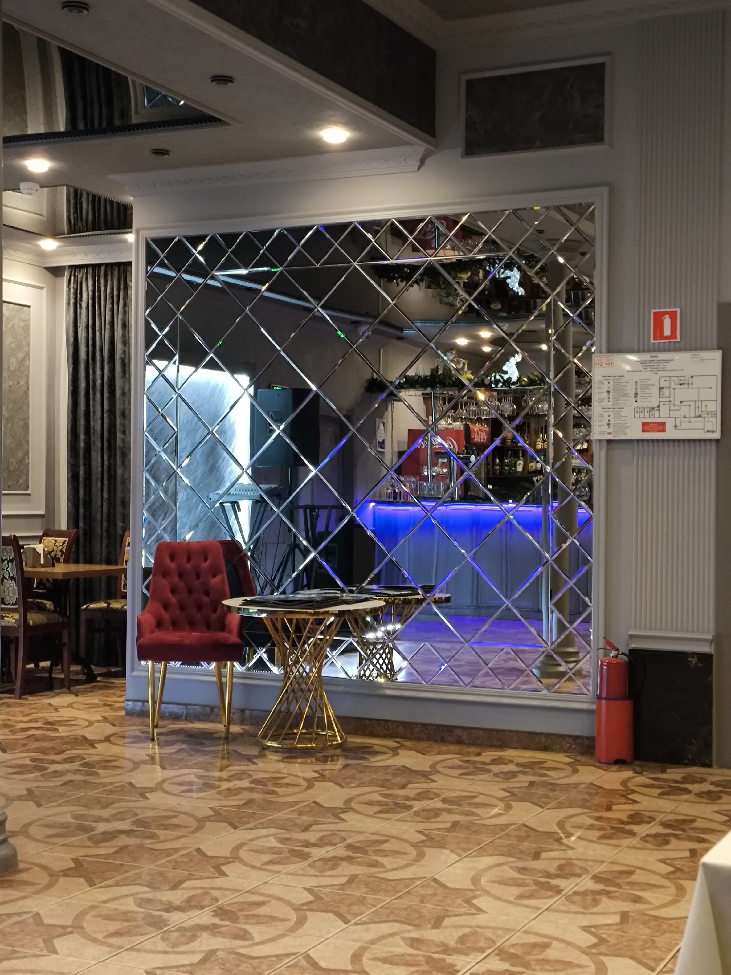 This image captures the interior of a sophisticated establishment, likely a restaurant, cafe, or hotel lounge, characterized by a mix of classic and modern design elements. The central focus is a large decorative mirror wall composed of numerous diamond-shaped beveled tiles, which reflects the opposite side of the room.
In the foreground, a plush red velvet armchair with elegant golden legs stands next to a small, round table featuring a glass top and an intricate golden twisted metal base. The floor is tiled with a repeating pattern of light brown and darker, star-like motifs. To the left, part of a dining area with wooden tables and chairs, partially obscured by dark grey patterned curtains, is visible. The ceiling is adorned with recessed lighting and decorative molding.
The mirror wall offers a reflection of a stylish bar area, illuminated with vibrant blue LED lighting under the counter. Shelves behind the bar display various bottles and glassware, suggesting a well-stocked liquor selection. Above the bar, more glasses are hanging, and some festive green decorations are visible, possibly holiday-themed. Also reflected are parts of the ceiling, additional lighting, and what might be sound equipment or a speaker.
On the right side of the image, mounted on a light grey fluted wall, is a white fire safety plan or evacuation map with a red square symbol indicating a fire extinguisher location. Below it, a bright red fire extinguisher is visible near the floor. The text "112 101" and "ПЛАН" (Plan) are discernible on the safety map, along with unreadable smaller text and a diagram. The ambiance is one of evening or night due to the reliance on artificial lighting, particularly the blue glow from the bar. No people are present in the scene. The location is specified as Khimki, Russia.