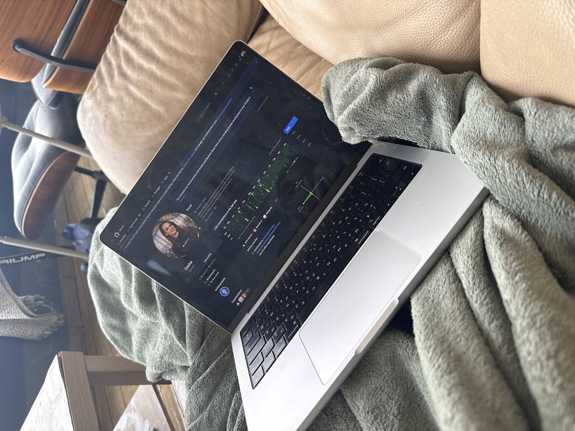 Here is a description of the image:
The image shows a laptop resting on a gray fuzzy blanket. The laptop screen displays a GitHub profile page with the name "Dasha" and the username "daxxac". It indicates the user is located in Tel Aviv, Israel. The laptop keyboard has Hebrew lettering. Other items in the background include a tan leather couch, a chair with a wooden seat, and what appears to be a box or container on the floor. The scene appears to be indoors.