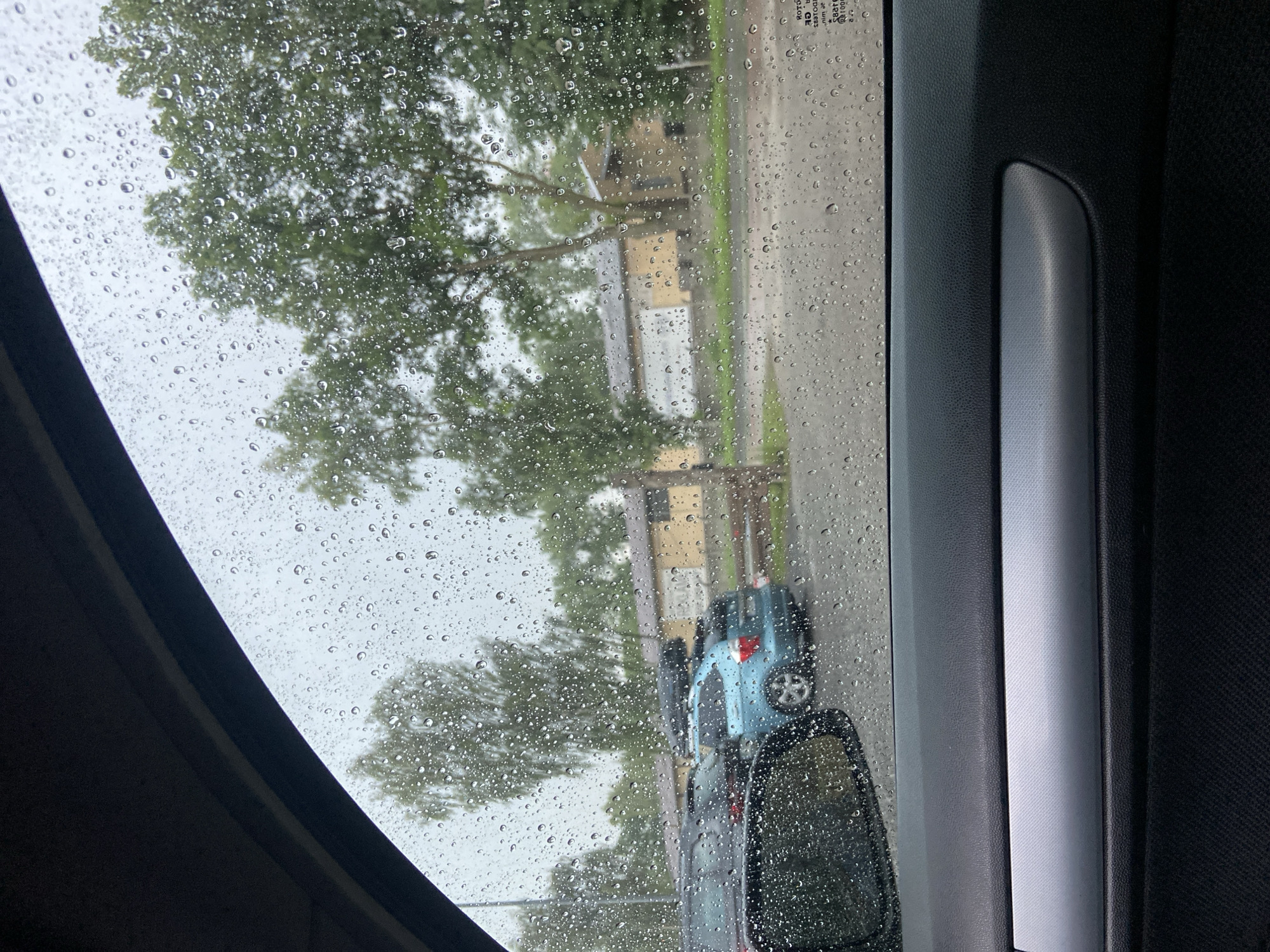 The image, taken from inside a car looking out through a rain-streaked window, depicts a parking lot on a cloudy day. In the foreground, the interior of the car is visible, including the door panel with a textured armrest and a metallic trim. The car's side mirror is also partially visible, reflecting the overcast sky.
Outside the car, water droplets distort the view of a row of parked vehicles. A blue SUV with a roof box is prominent, situated behind a darker vehicle. Beyond the cars, a light-colored building with a brown roof extends across the scene. Trees with dense green foliage are scattered throughout the background, suggesting a park or a wooded area adjacent to the parking lot. The overall atmosphere is somber, characterized by the rain and muted light.
The building in the background, with its distinctive architecture and the surrounding environment, strongly suggests a location within the city of Bolesławiec, Poland. This is based on the visual cues commonly associated with such buildings and landscapes in this specific region.