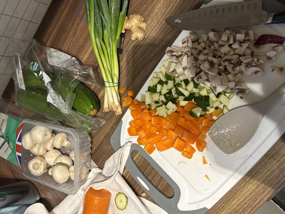 A wooden surface displays various ingredients prepared for cooking. Chopped carrots, zucchini, and mushrooms sit on a white cutting board next to a spoon. Nearby, are green onions tied with a blue rubber band, ginger, zucchini in a plastic bag, and packaged button mushrooms. A knife lies along the upper right edge. A kitchen towel sits beside a carrot and zucchini slice.