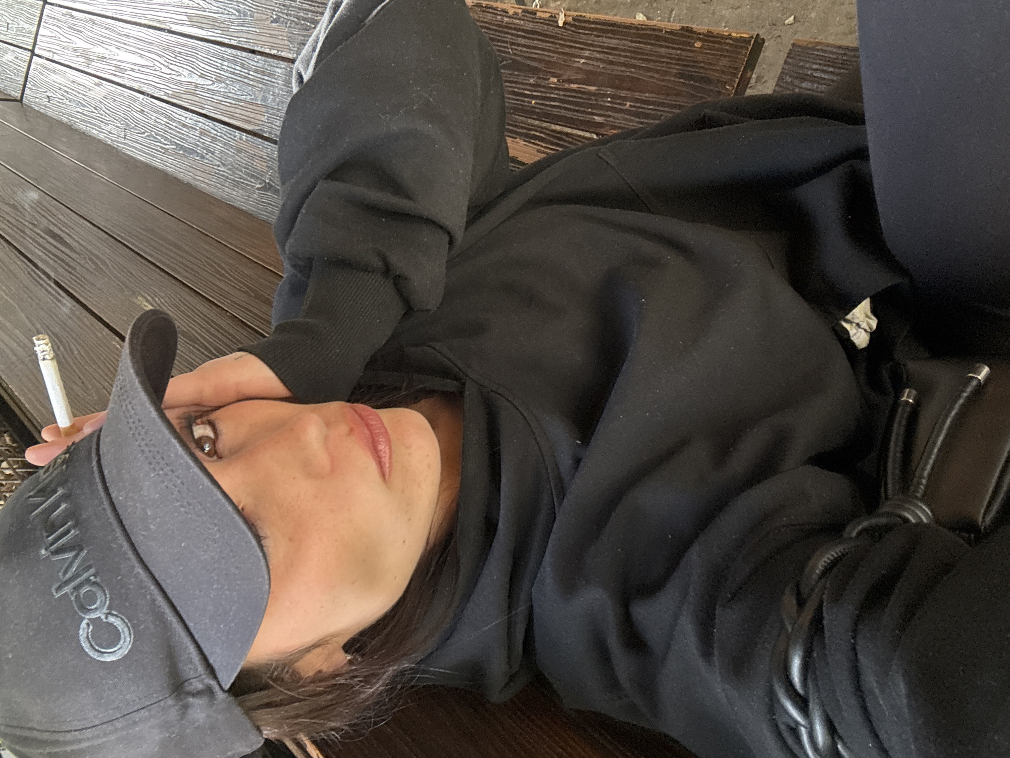 A person is lying on a wooden surface, possibly a bench or deck, with horizontal planks. The person is wearing a dark hooded sweatshirt and dark leggings or pants. A dark Calvin Klein baseball cap is on their head. They are holding a lit cigarette in their left hand. A dark purse or bag is visible near their right side. The location of the photo is not determinable from the image provided.