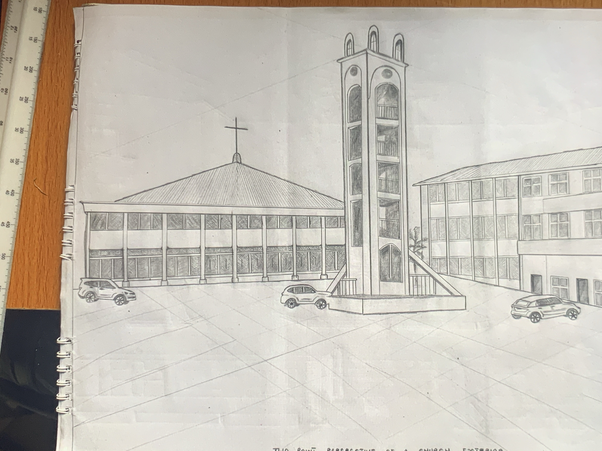 A pencil drawing of an architectural exterior scene is depicted on a sheet of white paper from a spiral-bound sketchbook. A white ruler with black numerical markings is visible along the left edge of the image, adjacent to the sketchbook's binding.
The drawing showcases a church complex, executed in a two-point perspective. The central structure is a church building with a large, multi-faceted, possibly pyramidal roof topped with a cross. Its facade features numerous rectangular windows with hatched shading. To the right of the church stands a tall, ornate bell tower with multiple levels, arched openings, and balconies. Further to the right, another multi-story building, possibly an annex or school, is shown with many windows, indicating a more utilitarian architectural style.
Three cars are parked in what appears to be a paved courtyard or parking area in front of the buildings. The cars are drawn with noticeable detail for their bodies and wheels. Faint grid lines are visible on the ground, common in perspective drawings, indicating the artist's use of a technical drawing method. The visible text at the bottom right of the drawing states, "TWO POINT PERSPECTIVE OF A CHURCH EXTERIOR." There is no activity taking place, as it is a static architectural illustration.