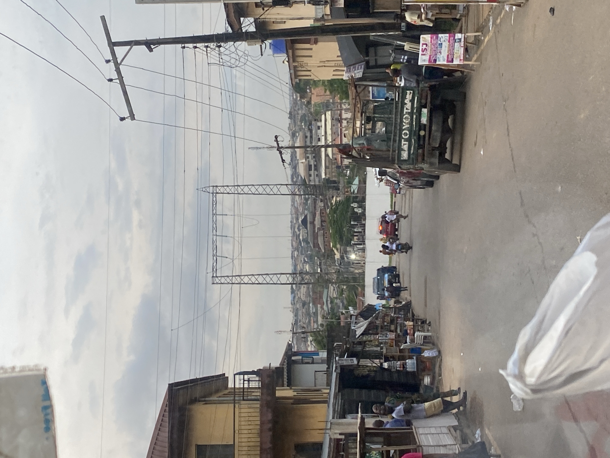 The image shows a street scene in Ikorodu, Nigeria, with buildings lining both sides of the road and a dense urban landscape in the background. In the foreground, a white plastic bag is partially visible, obscuring the lower portion of the frame. The street is paved and appears to be a busy thoroughfare with vehicles and people.
On the left side of the street, there are several low-rise buildings with shops at ground level. People are standing outside these shops, some interacting with each other. One building has a balcony with a railing. Overhead, power lines crisscross the sky, supported by wooden and metal poles.
On the right side of the street, more buildings are visible, also with shops. A red pickup truck with "TOYOTA" written on its side is parked. A sign for "ISJ" is displayed on an easel. People are also present on this side of the street, including individuals standing near the shops and a person on a motorcycle.
In the middle ground, vehicles are moving along the street, including a blue vehicle and a motorcycle. The background is filled with a panorama of houses and buildings stretching up a hill, indicating a densely populated area. Large electricity pylons are prominent in the distance, connected by thick cables. The sky is overcast with clouds.