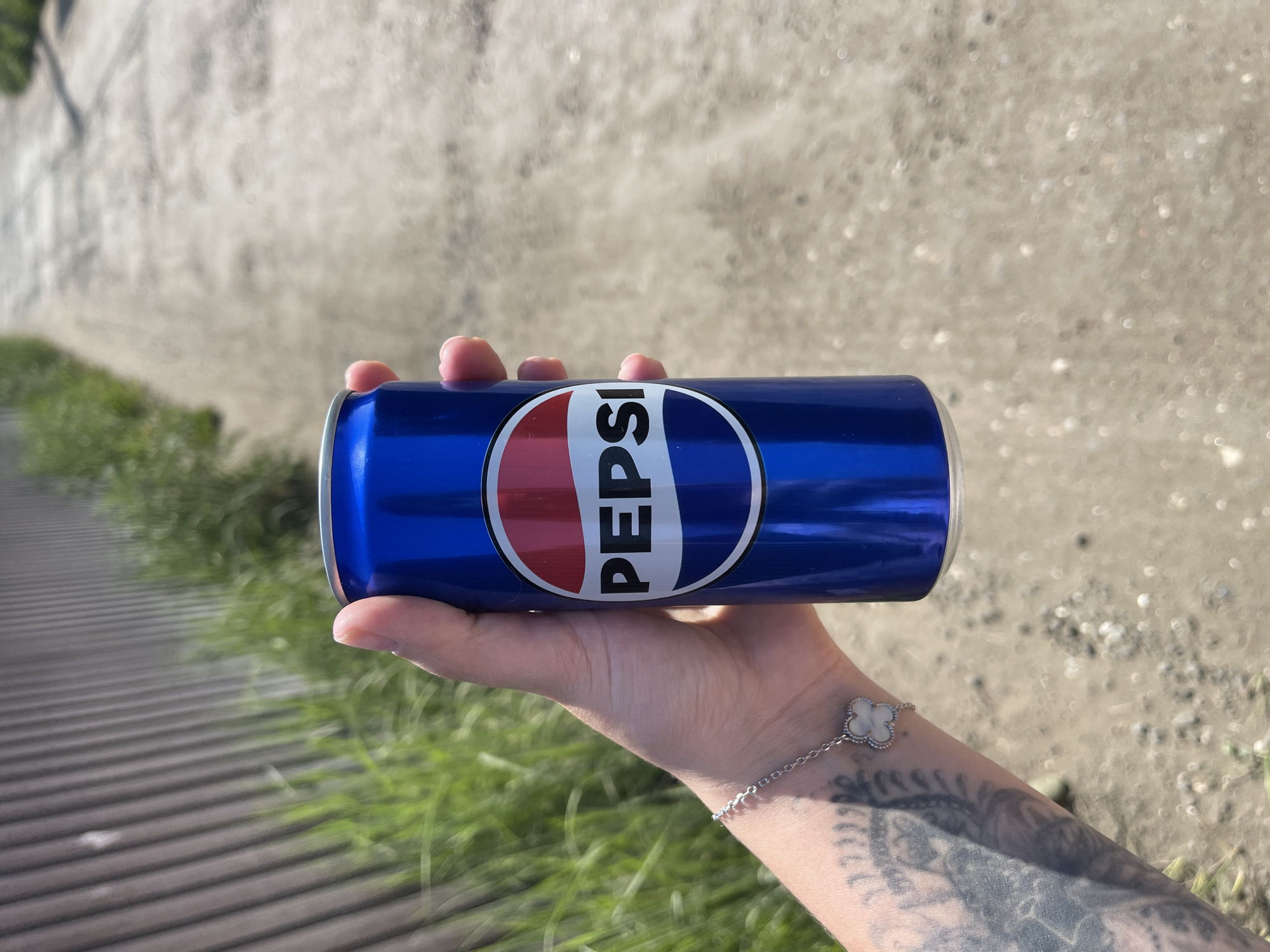 A person's hand holds a blue Pepsi can against a blurred background of a dirt path and a wooden fence. The Pepsi logo is clearly visible on the can. The person is wearing a bracelet with a clover-shaped charm. Tattoos are visible on the person's arm. The scene appears to be outdoors, possibly in a rural or suburban setting. No specific location in Molokovo, Russia, can be determined from the image.