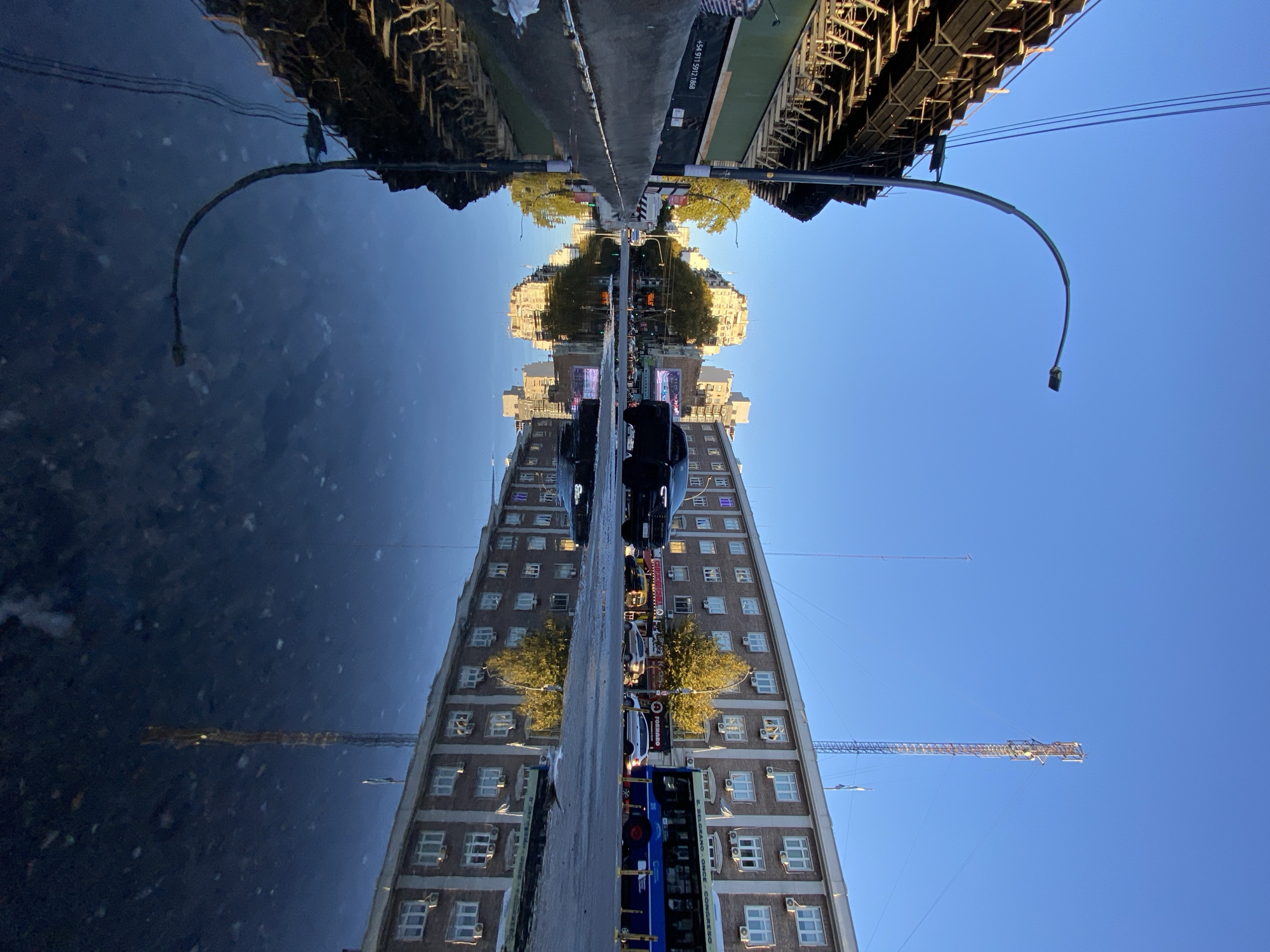 The image shows an upside-down view of a street in Buenos Aires, Argentina, reflected in water. The primary subject is a tall brick building with numerous windows, aligned vertically along the right edge of the image. There are trees, parked cars, and a bus along the street in front of the building. A crane is visible at the top right corner. Above the reflection, there is a road flanked by structures that appear to be bridges or overpasses, with streetlights curving downwards towards the center. The sky is clear and blue. A text that says "+54 911 5971 21668" is visible along the road. The image is a street scene reflected on the street water.