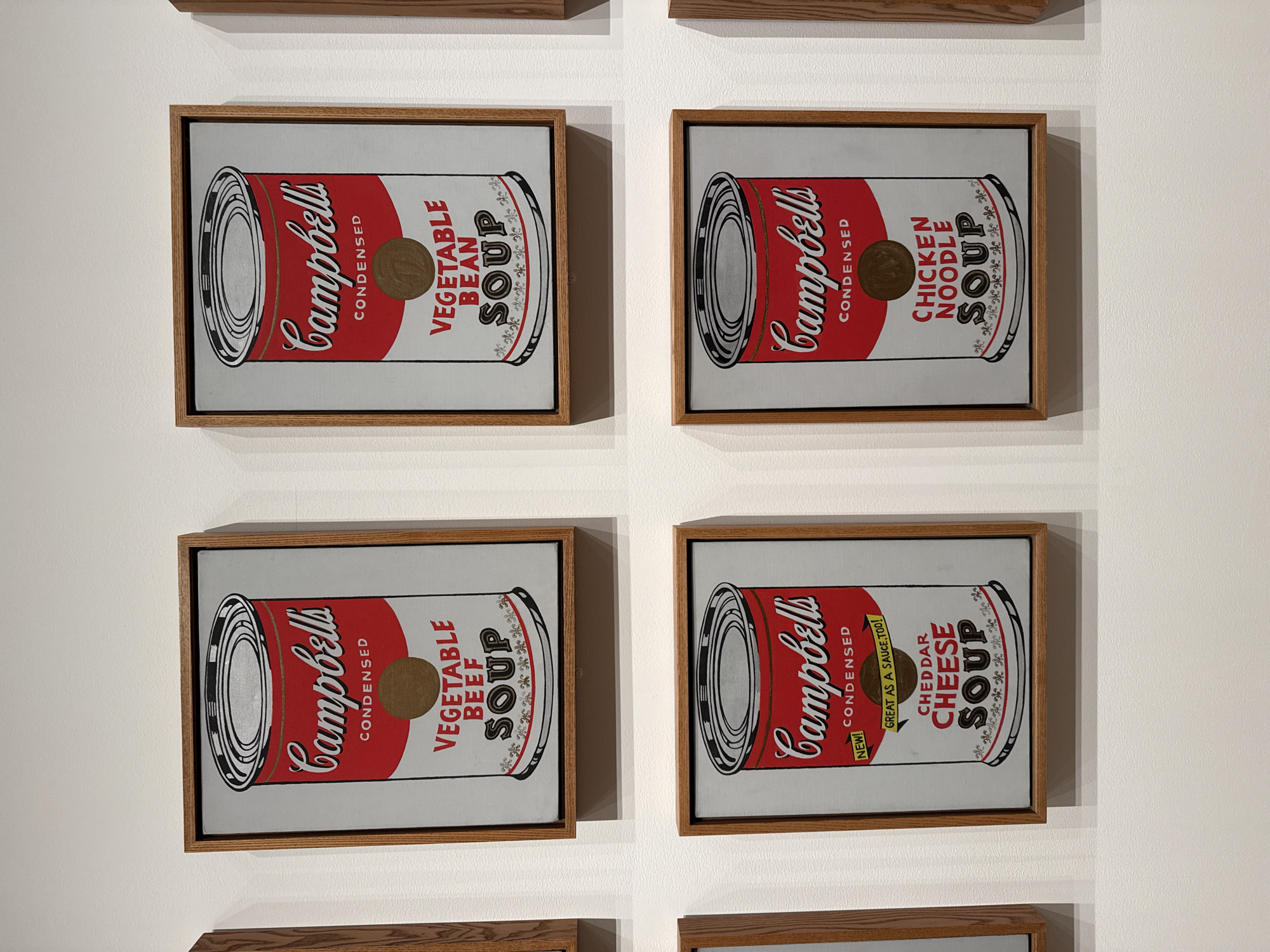 The image shows a display of four framed pop art prints depicting Campbell's soup cans, a work reminiscent of Andy Warhol's iconic series. Each print features a different variety of soup: Vegetable Bean, Chicken Noodle, Vegetable Beef, and Cheddar Cheese. The cans are rendered in a stylized, flat design with red and white coloring. The frames are a light-colored wood. The background is a plain white wall. The exact location cannot be determined from the image but is likely within a gallery or museum setting.