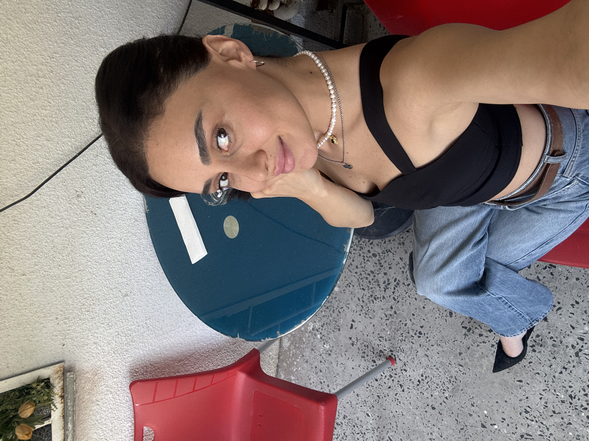 An outdoor scene is depicted, likely a patio or balcony, with a woman seated. She has dark hair pulled back and is wearing a black wrap-around top, light blue jeans, and black pointed-toe heels. She is adorned with a pearl necklace and a delicate pendant. Her left hand rests on her cheek as she looks towards the camera with a slight smile.
To her left, a red plastic chair is visible, and in front of her, a round, teal-colored table with a white piece of paper on it. The ground is a terrazzo-style concrete. In the background, a textured white wall features a rustic framed object with some artificial flowers. A black cable runs diagonally across the wall.
Given the style of the architecture and the common presence of such outdoor seating areas in residential and commercial establishments, this scene could be located in the Vake or Saburtalo district of Tbilisi, Georgia, areas known for their modern apartment buildings with balconies and patios.