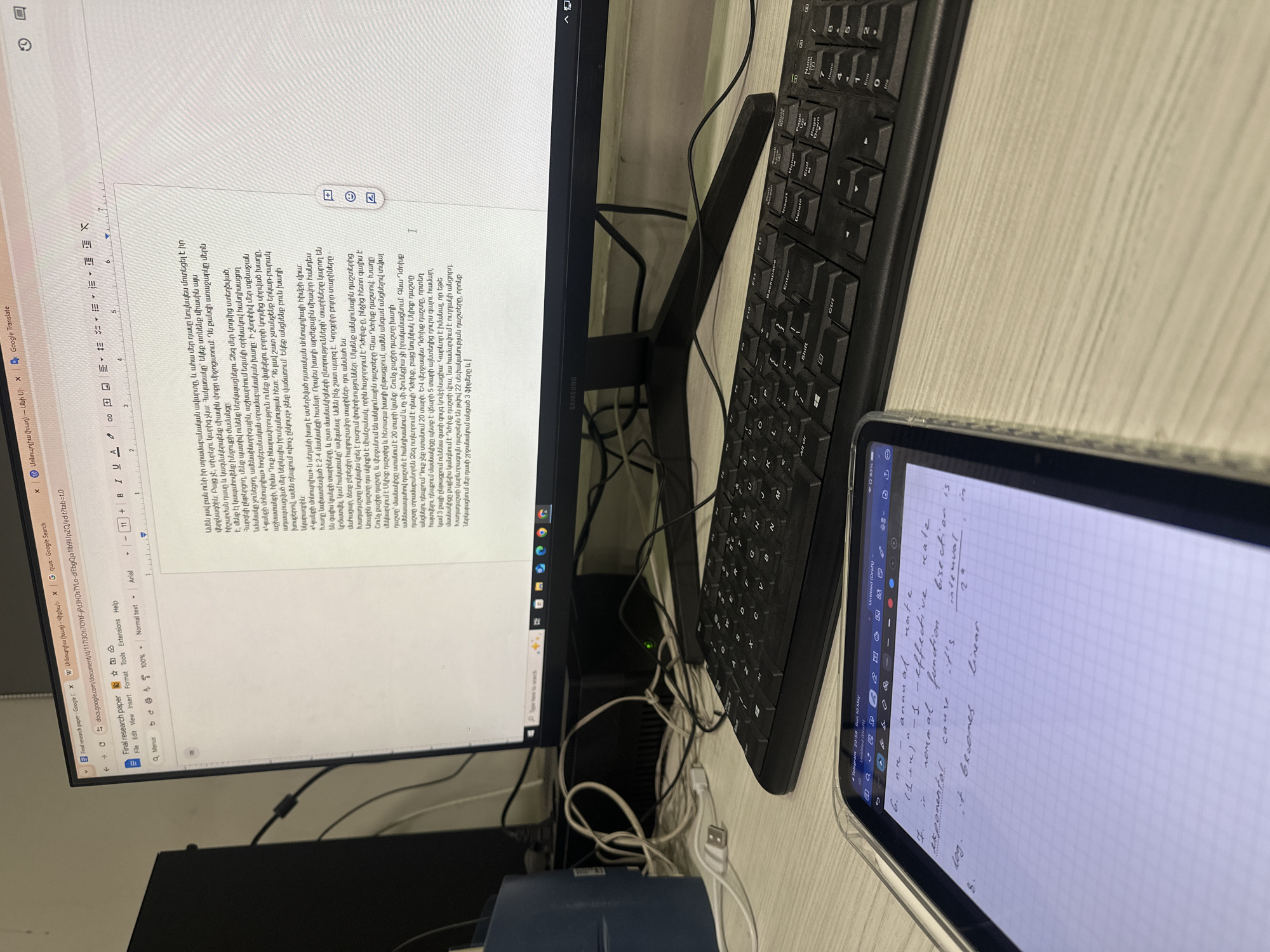 The image shows a desk setup with a monitor, keyboard, and tablet. The monitor displays a Google Docs document in Armenian, titled "Final research paper." The keyboard is black and standard. The tablet shows handwritten notes on a grid-lined page, containing mathematical-looking formulas and terms like "annual rate," "effective rate," "bisection," and "linear." The desk surface is light-colored and appears to be made of wood or a similar material. The scene is cluttered with cables and wires. A dark object is beneath the monitor, near the cables. The exact location, city, or country is not evident from the image.