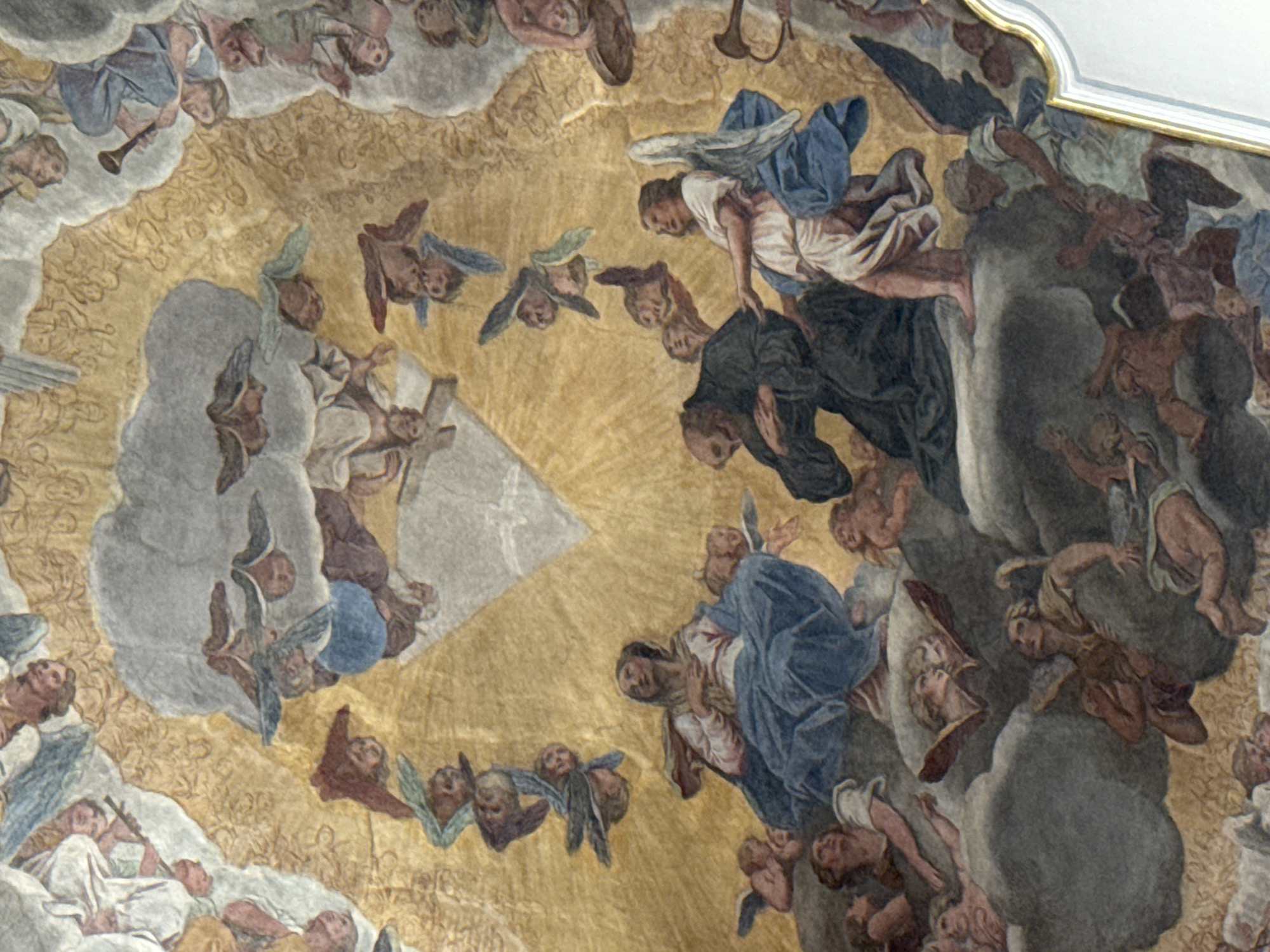 The image depicts a fresco of a celestial scene, likely a religious painting intended for a church or cathedral ceiling. The central figures are a woman seated on clouds, possibly the Virgin Mary, draped in a blue mantle and white garment, and a robed male figure kneeling before her. To the right of the kneeling figure is an angel with blue wings, reaching out to the man. Numerous other figures, appearing to be angels or cherubs, are scattered throughout the composition, some with wings, some playing instruments, and others ascending or descending. They are rendered in a dynamic and ethereal manner, surrounded by clouds and beams of golden light emanating from a triangular form at the apex of the fresco, suggestive of divine presence. The overall impression is one of ascension and divine revelation.
This fresco is located in the ceiling of the Jesuit Church (Kirche St. Franz Xaver) in Lucerne, Switzerland.