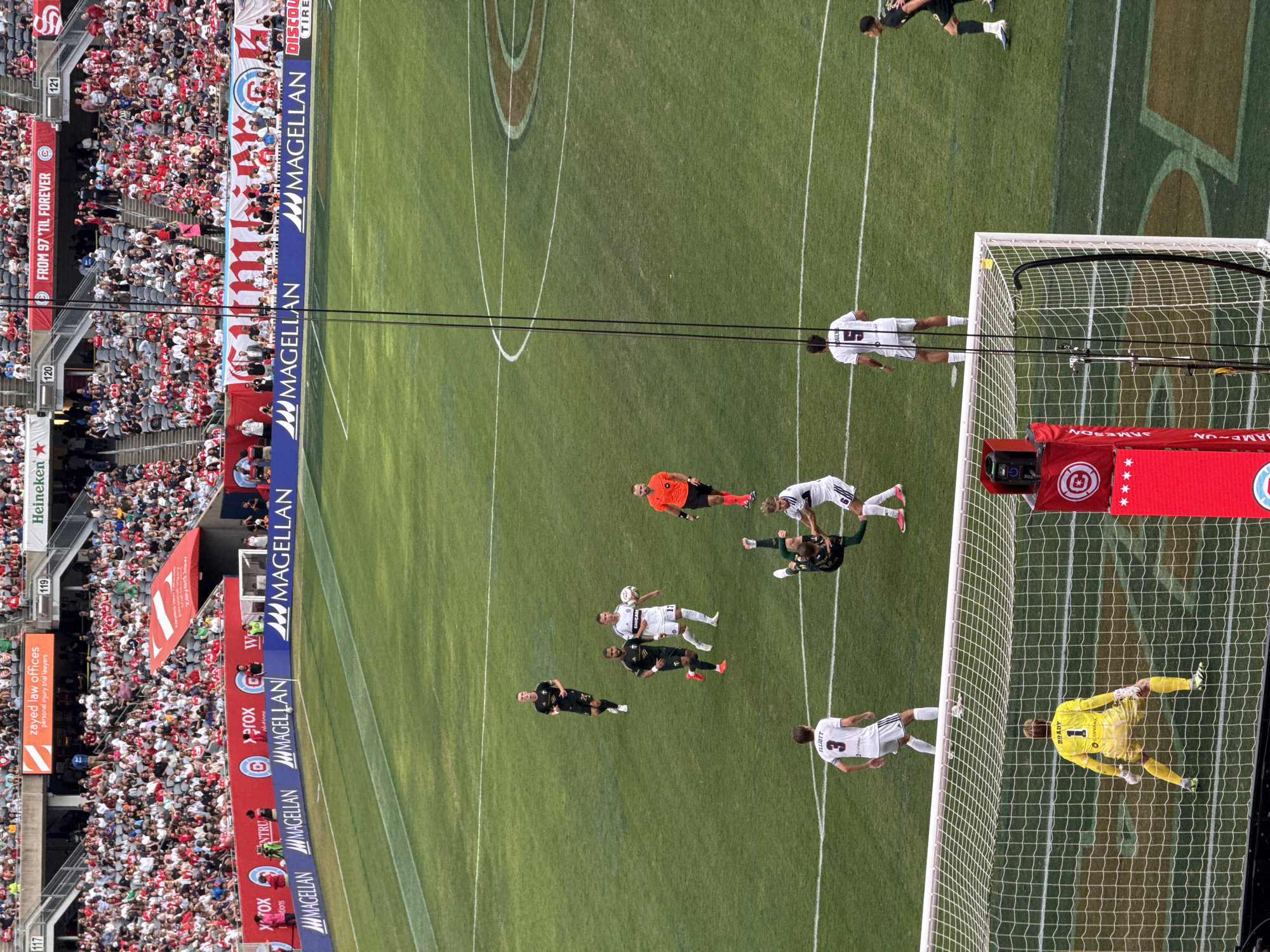 The image shows a soccer game in progress at Soldier Field, located in Chicago, Illinois, United States of America. The stadium is filled with spectators in the stands, many wearing red and white. On the field, several players are visible. Two players in white uniforms are near the center circle, one with the ball at his feet, and another in a black and green uniform is engaged in a sliding tackle on a player in a white and green uniform. A referee in an orange shirt is also present. In the foreground, a goalkeeper in a yellow uniform is positioned in front of a soccer goal. The field is marked with white lines, and there are advertisements visible on the perimeter of the field and on the stadium structure.