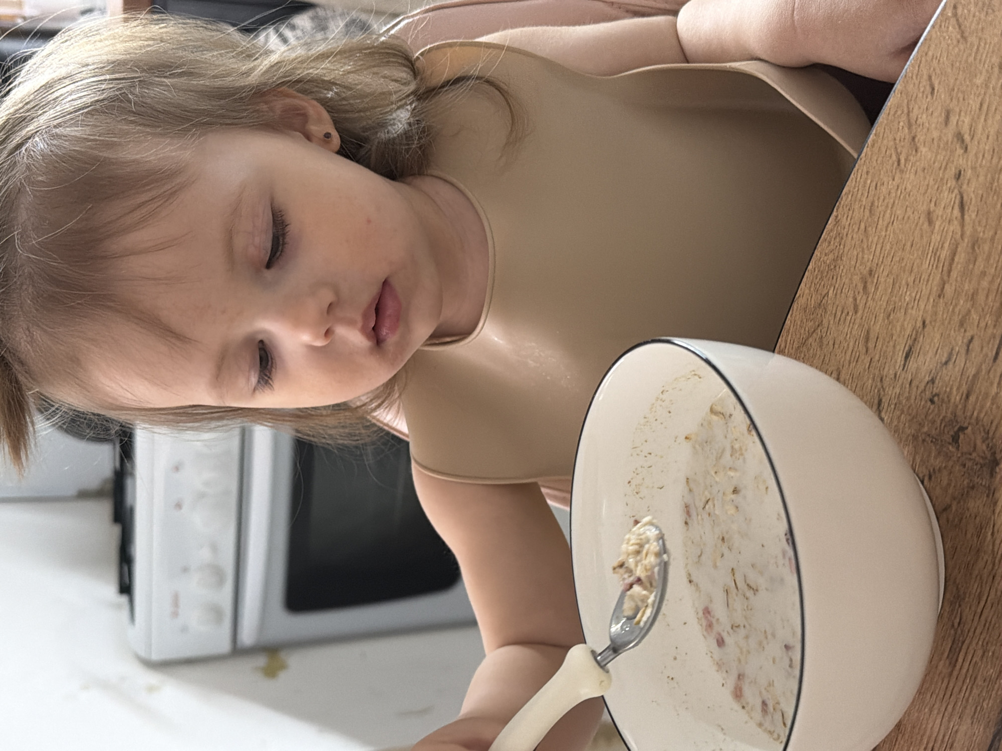 A young child is seated at a wooden table, wearing a beige bib, with a white bowl of partially eaten cereal or oatmeal in front of her. She is holding a spoon with some of the cereal. The child appears to be looking down at the bowl. In the background, partially visible, is a white microwave oven. The scene is indoors. The location is not discernible from the image and there is no indication that it is in Reshetnikovo, Russia.