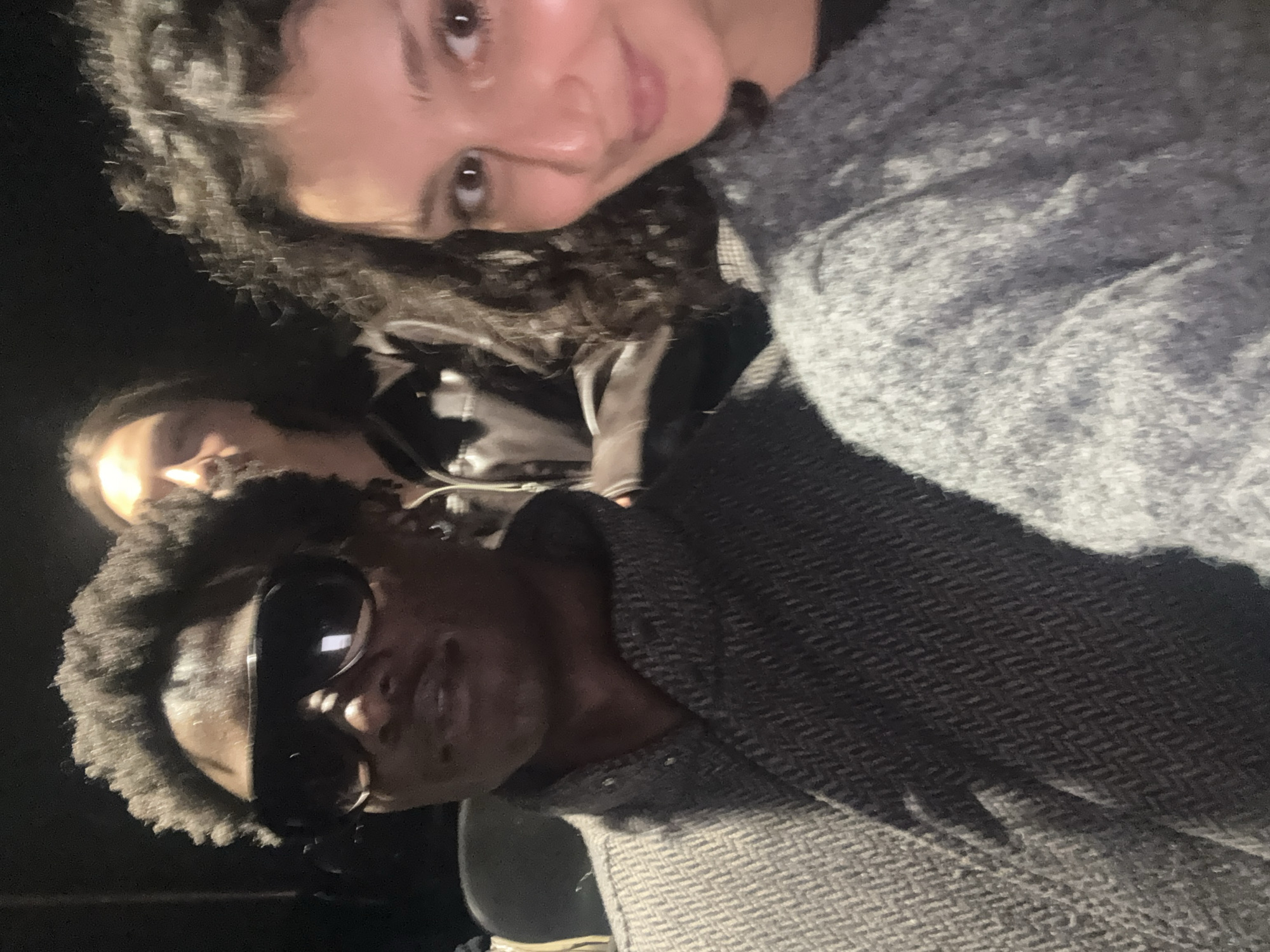 A selfie taken indoors at night features three people. In the foreground, a Black man with dark skin and short curly hair wears sunglasses and a gray herringbone coat. To the right, a White woman with dark curly hair and dark eyes is smiling. Behind them and to the left, another person with light skin is visible. The location is presumed to be in the city of London, United Kingdom, but lacks visual confirmation in the image.