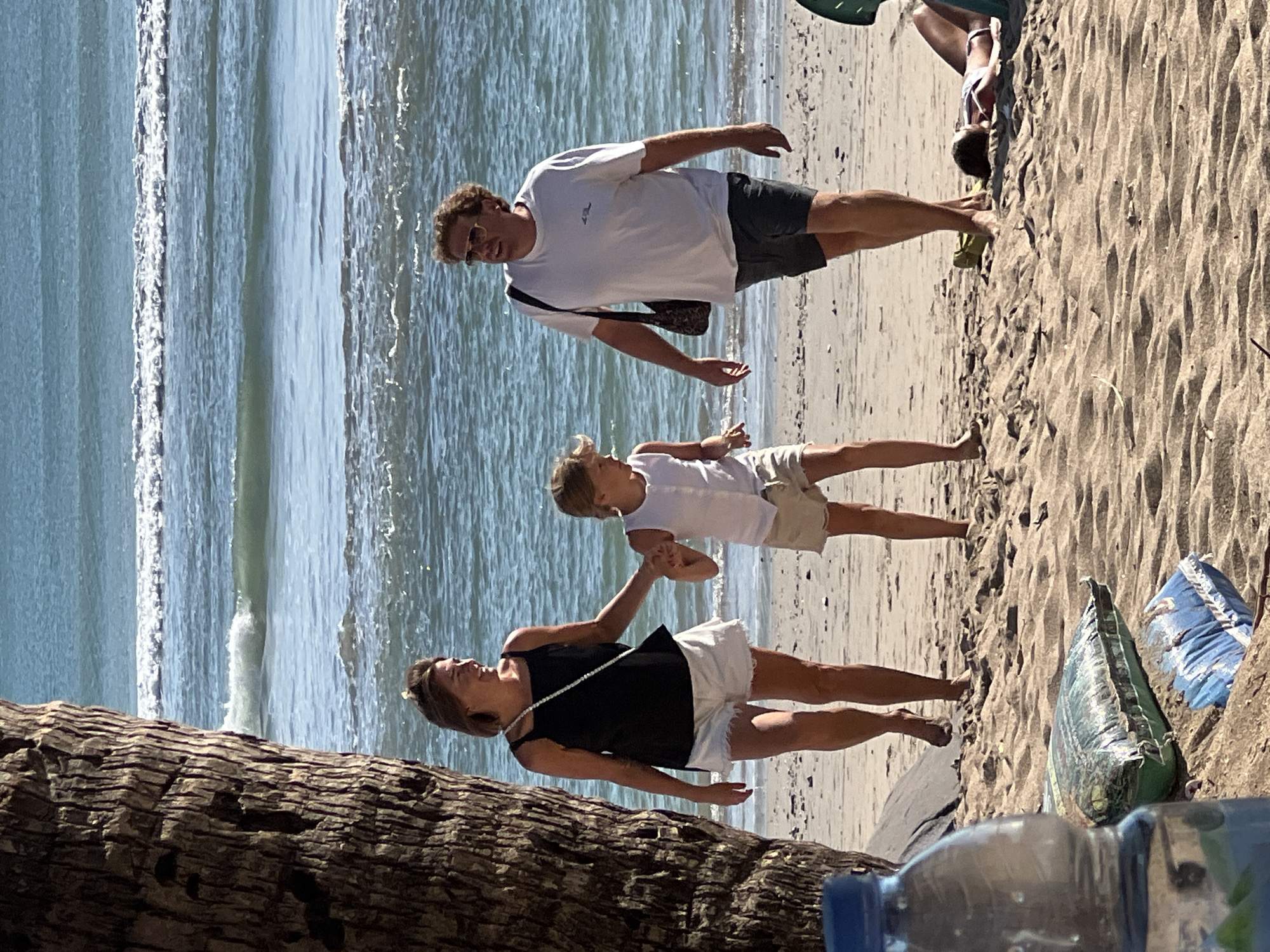 At Kuta Beach, Bali, Indonesia, on a sunny day, a family walks along the sandy shore. A woman in a black tank top and white shorts leads a child in a white tank top and beige shorts, holding the child's hand. A man in a white t-shirt and dark shorts follows them, carrying a bag over his shoulder. The ocean with gentle waves stretches out behind them. In the foreground, a plastic water bottle and some bags lie on the sand. Further back on the beach, another person is lying down, possibly sunbathing. The scene captures a relaxed vacation atmosphere.