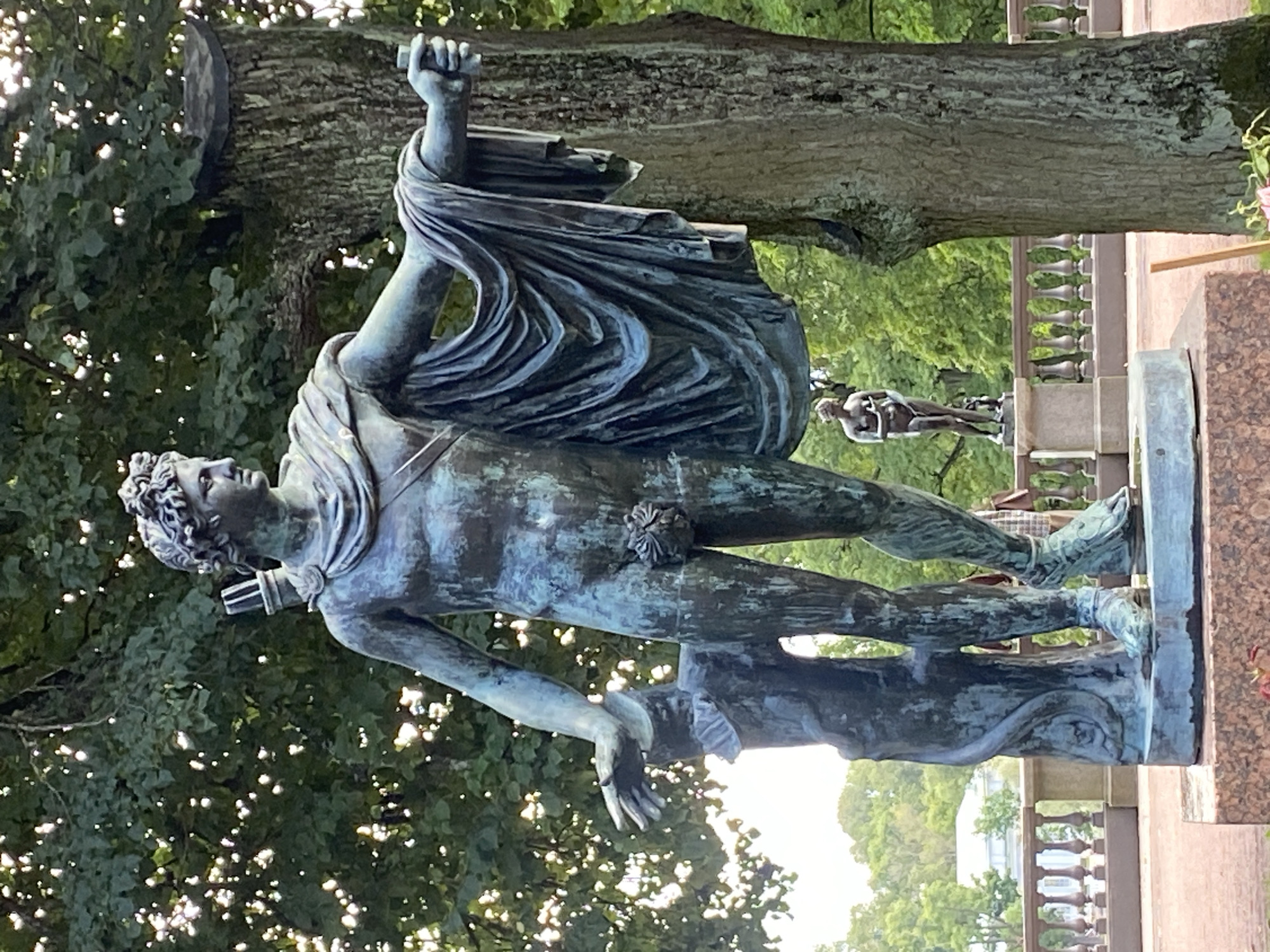 The image shows a full-body bronze statue of a male figure, identified as Apollo Belvedere, in a park setting. The statue is positioned in the foreground, with a large tree trunk to its right and lush green foliage behind it. The statue is depicted in a contrapposto stance, with his right arm extended and his left arm bent, resting on a tree stump. He wears a draped cloth and has a quiver of arrows on his back. The bronze has a weathered, greenish patina.
In the background, a balustrade runs horizontally across the middle ground, separating the area where the main statue stands from a lower level. Beyond the balustrade, there is another, smaller statue of a female figure. Further in the distance, trees and buildings can be seen, suggesting a park or garden environment. The overall impression is one of classical statuary set within a natural landscape.
This statue is located in the Pavlovsk Park, near Saint Petersburg, Russia. Specifically, it is part of the collection of sculptures adorning the park's grounds, often found near prominent architectural features or scenic viewpoints. The park itself is renowned for its English landscape design and its collection of classical sculptures.