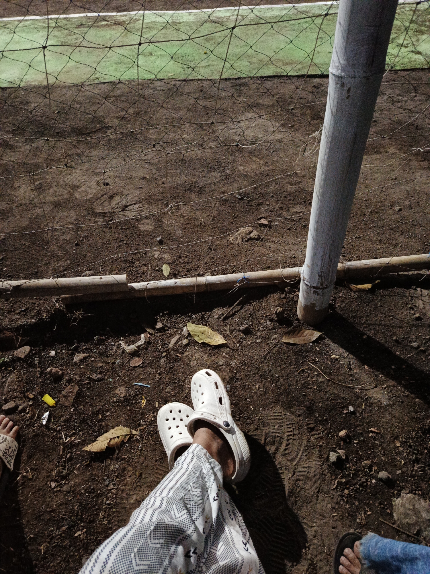 The image depicts a ground-level view of a dirt area near a sports field, likely a futsal court, in Rowokangkung, Indonesia. A section of the green sports field with a white boundary line is visible in the upper portion of the image, separated from the dirt area by a net fence. A thick bamboo pole, part of the fence structure, stands vertically on the right side, casting a shadow. A horizontal bamboo pole runs across the dirt area, secured by string. The ground is covered in dirt, small stones, and scattered dry leaves, with some debris including a yellow plastic wrapper and a piece of white plastic. Two individuals' legs are visible in the foreground. One person is wearing white Croc-style sandals and patterned pants, with their left leg extended. Another person's legs are partially visible on the far right, wearing dark sandals and blue jeans. The scene appears to be outdoors at dusk or night, given the lighting and shadows.