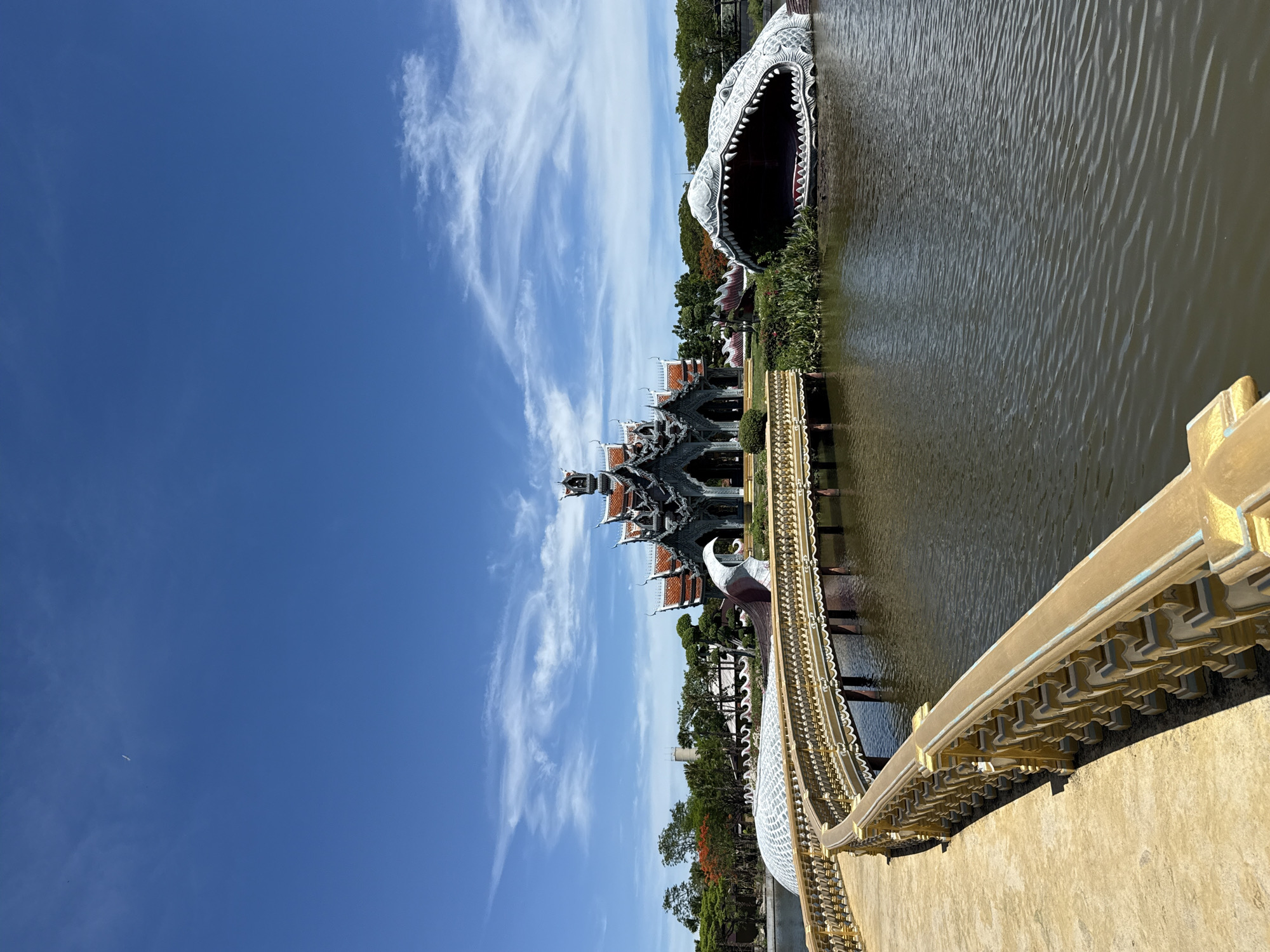 The image shows a view of the Ancient City (Muang Boran) in Bang Pu Subdistrict Municipality, Thailand. A large, open-mouthed crocodile sculpture is partially submerged in the water. A decorative Thai-style pavilion with layered roofs is visible in the background. A golden-colored bridge with detailed railings leads toward the pavilion. The sky is blue with scattered cirrus clouds. The water appears still. Vegetation lines the water's edge.