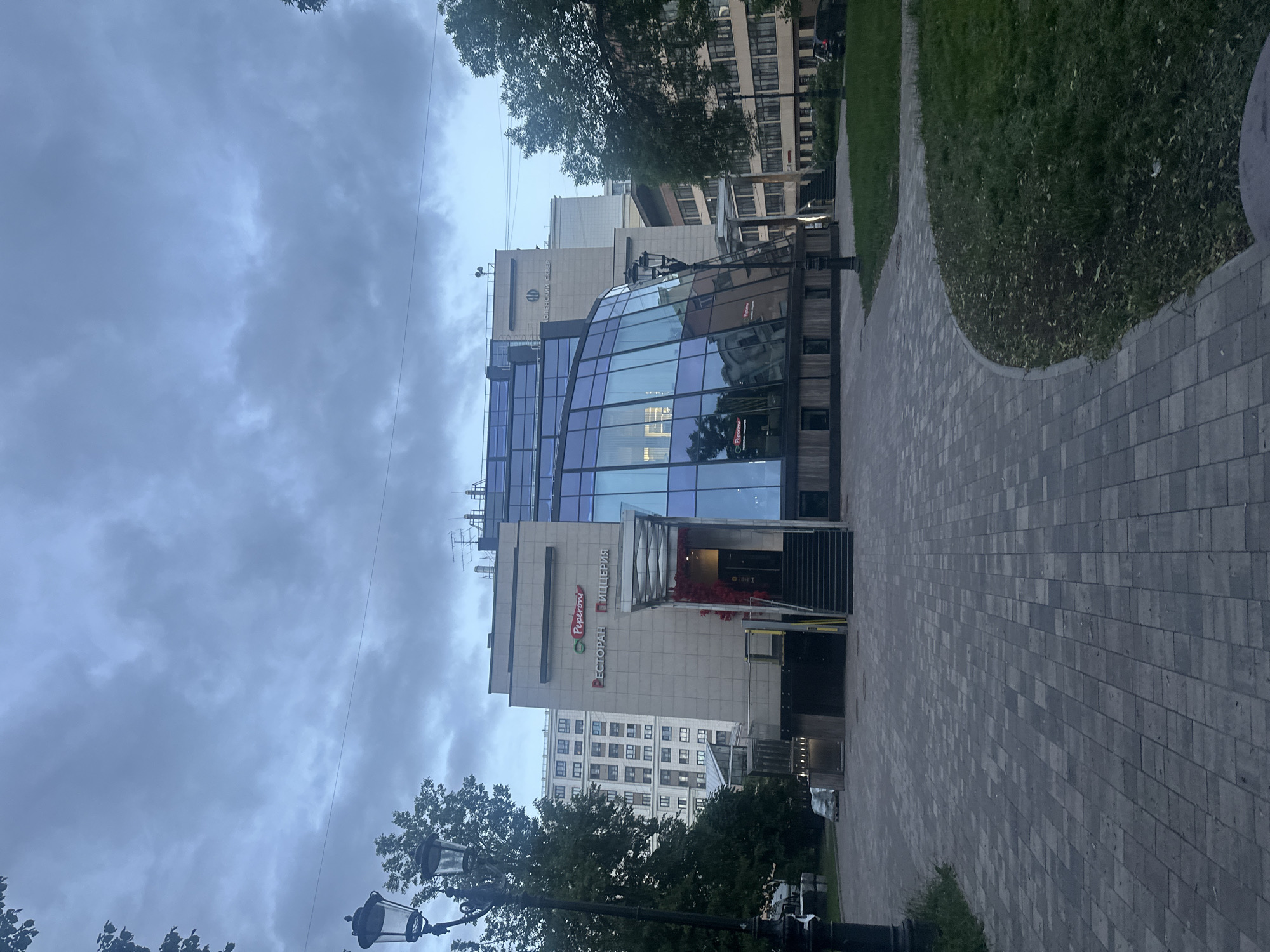 The image depicts a modern building with a distinctive curved glass facade, reflecting the overcast sky and surrounding structures. The building is situated in a city square with a paved walkway in the foreground, flanked by a grassy area on the right. A streetlamp with a classic design stands on the left side of the paved area.
The building's facade features signage that includes the word "Peperoni" in red lettering, along with Cyrillic text that appears to read "РЕСТОРАН" (Restaurant) and "ПИЦЦЕРИЯ" (Pizzeria). A glass-covered entrance with stairs leads up to the building's interior. The architecture suggests a contemporary commercial or public space.
In the background, other buildings are visible, including a tall, light-colored residential building with numerous windows and a beige building with Cyrillic lettering above the entrance. The overall atmosphere is subdued, likely due to the cloudy weather. No people are clearly visible in the scene, and there are no apparent interactions occurring. The environment appears to be a well-maintained urban space.
