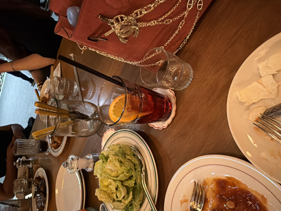 An overhead shot reveals a wooden table filled with dining accoutrements. There's a half-full red cocktail with an orange slice and straw, glasses with breadsticks, a plate of green salad, and various dishes with food remains, including a plate with a light-colored, flaky dish and another with a brownish sauce. A pink handbag with a gold chain is near the top of the frame. Utensils such as forks are scattered about. Portions of people are visible on the sides of the frame.