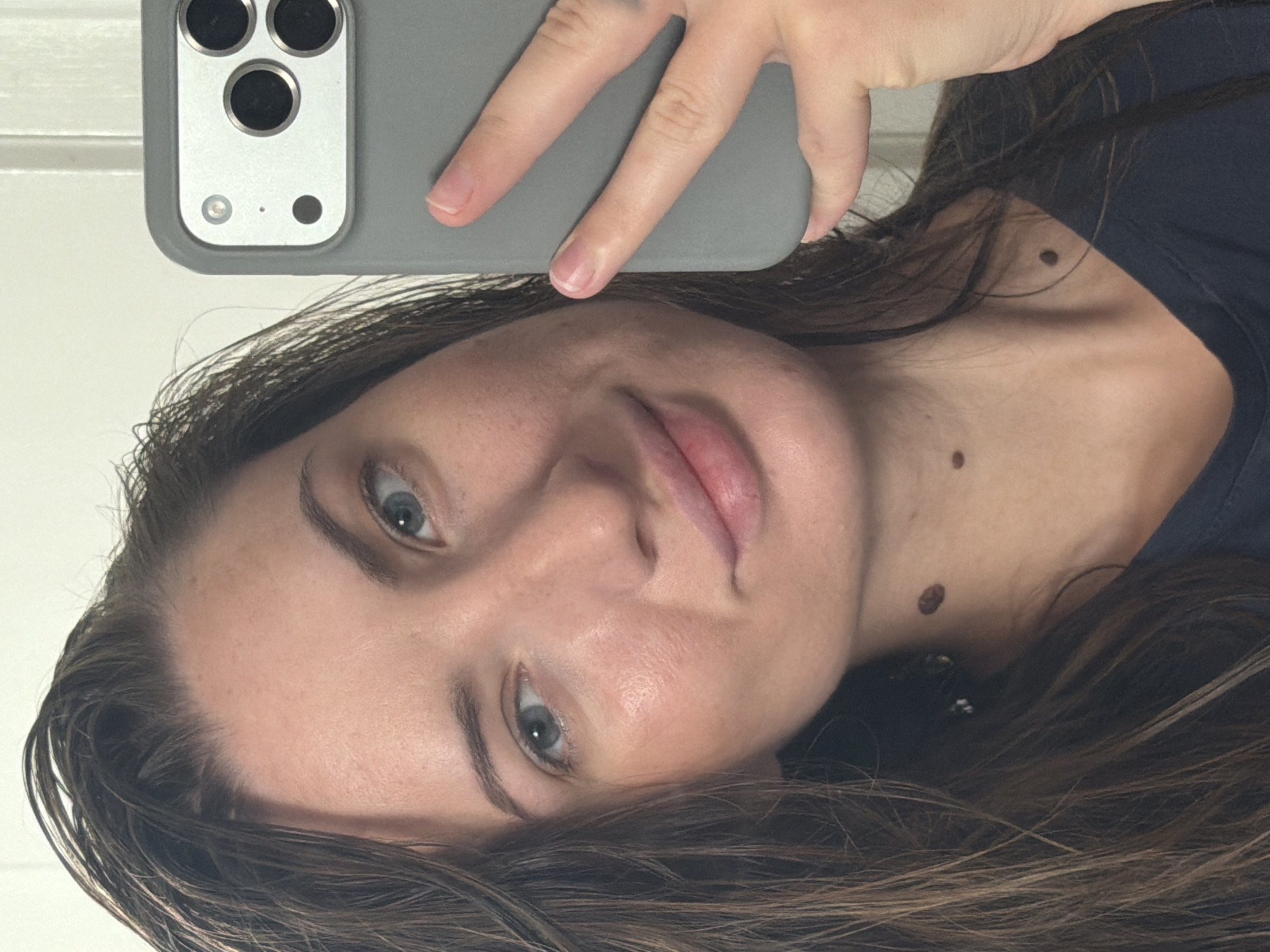 A woman with light skin, blue eyes, and long brown hair is taking a selfie in a bathroom. She is holding a gray phone with a triple camera array in her right hand. Her left hand is partially visible, resting on the phone. The woman is wearing a dark blue t-shirt. Her hair appears wet and is falling around her face and shoulders. There are several moles visible on her neck. The background is a white wall with a vertical panel. The image is a close-up shot, focusing on the woman's face and upper body.