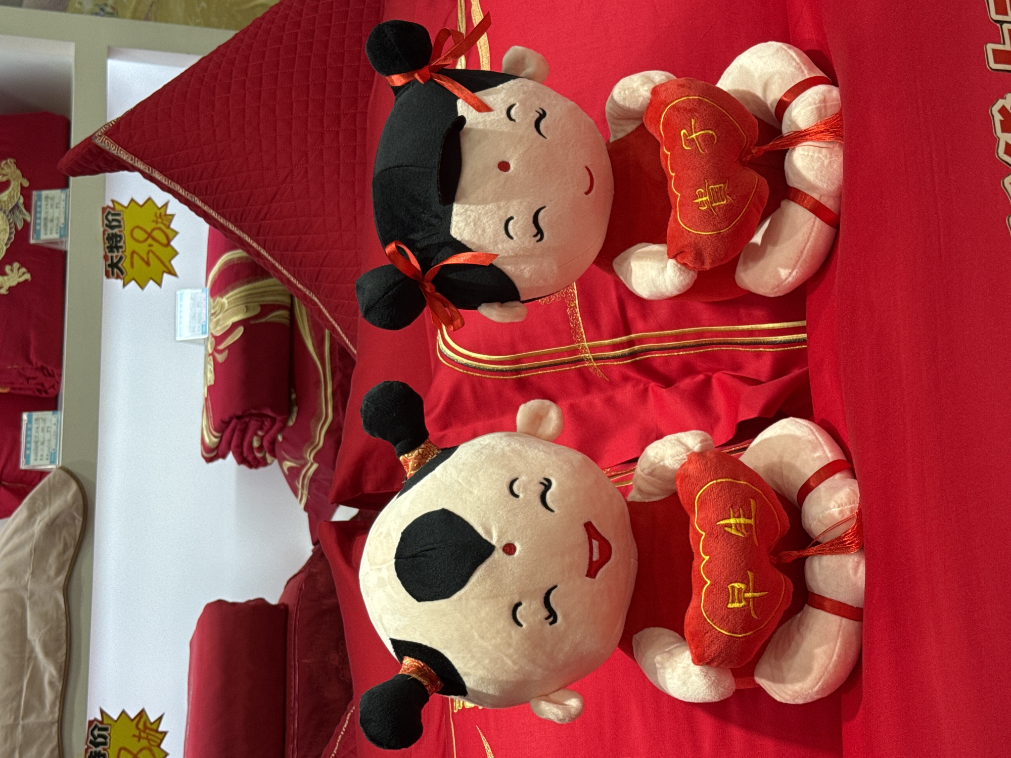 Two Chinese-style plush dolls lie on a vibrant red surface, likely fabric or bedding, in what appears to be a retail display. One doll has black hair styled in buns tied with red ribbons; the other doll has a white face with a black spot. Each doll has small embroidered features depicting a smiling face. Attached to each doll's back is a small red cushion with embroidered gold-colored Chinese characters and white tubular fabric sections with red ribbons, resembling a lucky charm. In the background, there is a display of red bedding, and a price tag with Chinese characters is visible. The scene may be located in a retail establishment in Qianjiang, China, given the style of the dolls and other bedding, which are often associated with Chinese culture and festivities.