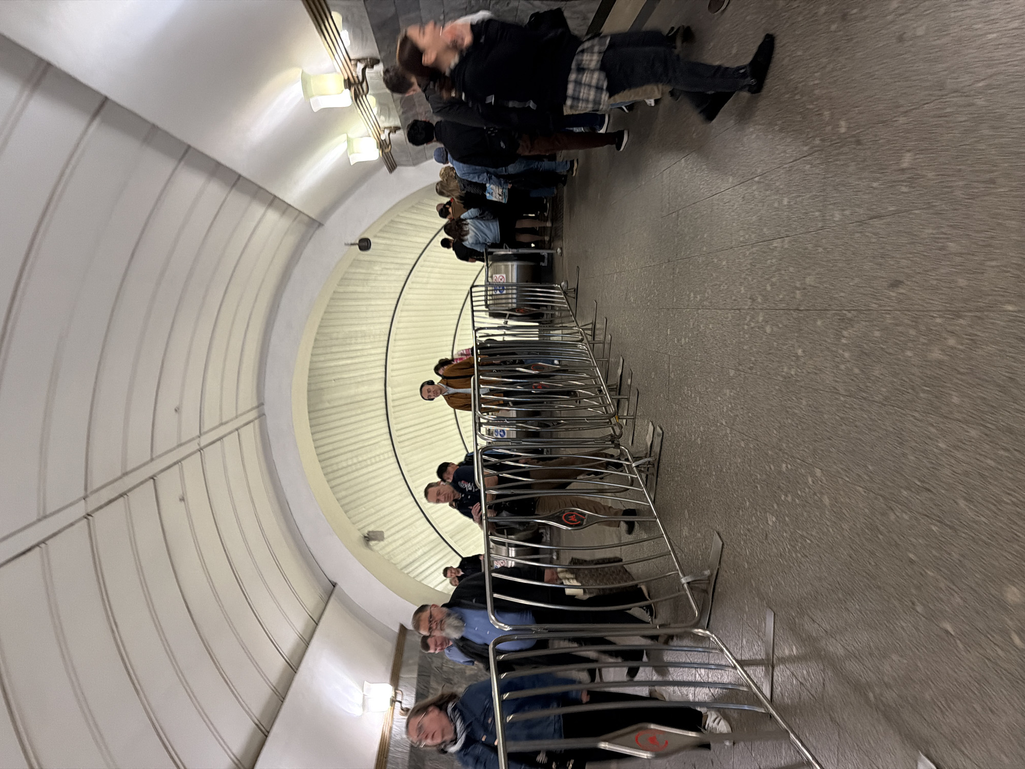 A crowd of people are walking through an underground passageway. The passageway is lit by overhead lamps and has a curved ceiling. Metal barricades are set up to guide the flow of people. The floor is paved with a speckled material. The people are dressed in casual clothing and appear to be of various ages and genders. Some are walking in groups, while others are alone. The overall impression is one of orderly movement and transit.