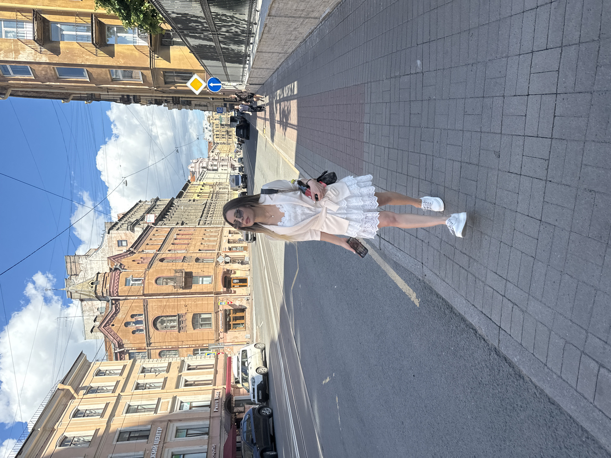 A young woman stands on a paved sidewalk next to a street in Saint Petersburg, Russia. She is wearing a white lace dress, a light pink cardigan, white sneakers, and sunglasses. She is holding a smartphone in her right hand and a red object, possibly a phone or wallet, in her left. The scene depicts a sunny day with blue skies and fluffy white clouds.
The street is lined with traditional European-style buildings with ornate facades and multiple windows. Cars are parked along the street and a tram track runs down the middle. Traffic signs are visible, including a yellow diamond sign and a blue circular sign with an arrow. The architecture suggests an urban setting in Saint Petersburg. The woman appears relaxed and is looking towards the camera. The time of day appears to be daytime, with clear sunlight casting shadows on the sidewalk.
There is visible text on signs on the buildings. One sign reads "Ринто Бак", another partially visible sign says "ский центр". The overall atmosphere is one of a pleasant day exploring a historic city.