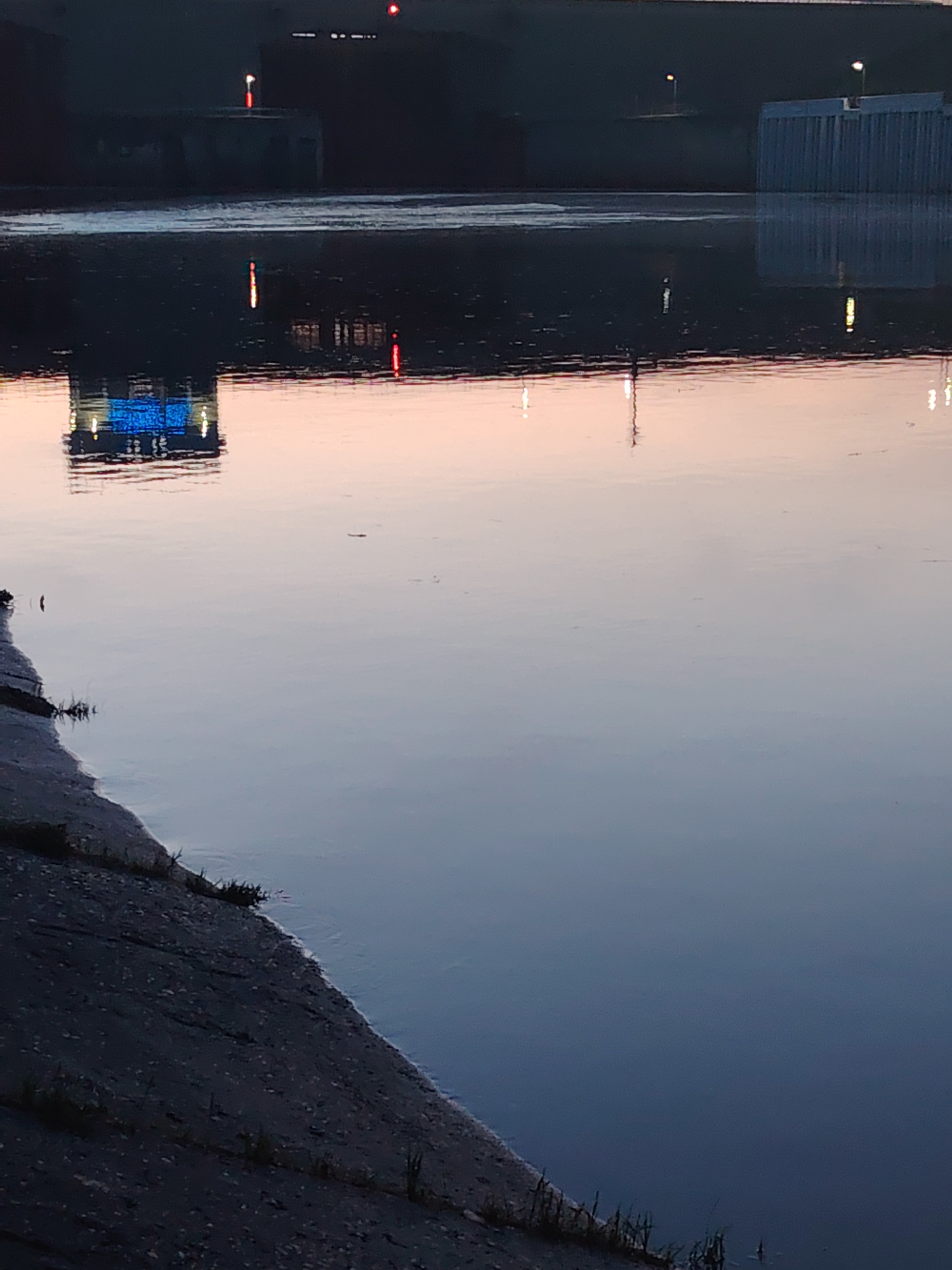 The image depicts a body of water at dusk or dawn in Chaikovsky, Russia. The water is smooth, reflecting lights and structures from the far side. A concrete embankment is visible in the foreground, with sparse vegetation growing along the edge. In the distance, there are structures that appear to be part of a dam or industrial facility, some with lights. The reflections in the water create an abstract and mirrored image of the distant scene.