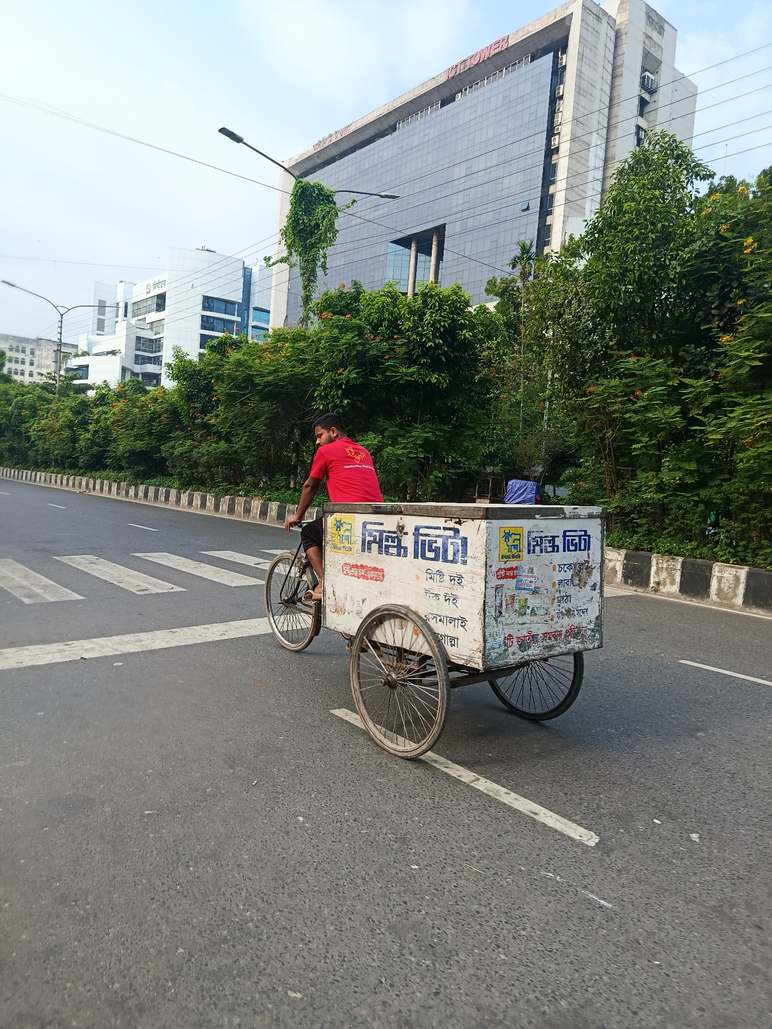 The image depicts a man riding a tricycle on a road with a crosswalk in what appears to be a city environment. The man is wearing a red shirt and dark shorts. The tricycle has a white container attached to the back, covered with writings in Bengali language. The buildings in the background include a structure labeled "ICT TOWER". The location is Dhaka, Bangladesh.