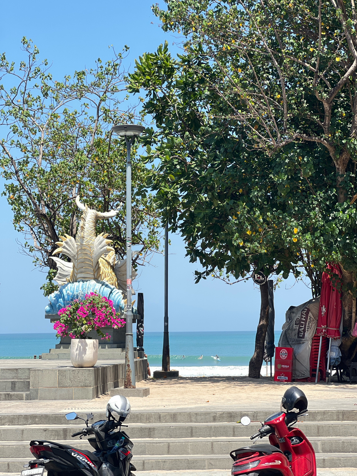 This image depicts a scene in Kuta, Indonesia, likely near the beach. In the foreground, two motorbikes are parked on a paved walkway adjacent to concrete stairs. The motorbike on the left is black with red accents, and the one on the right is red. Both have helmets resting on their handlebars. Behind the motorbikes, a tiered concrete structure rises, leading towards the beach.
On the left side of this structure, a white pot overflowing with vibrant pink bougainvillea flowers sits prominently. Behind the flowers is a large, ornate statue of a mythical creature, possibly a dragon or a sea serpent, with white and gold detailing, resting on a blue, wave-like base. A street lamp stands to the right of the statue.
Beyond the concrete structure, a sandy beach stretches out, meeting the turquoise waters of the ocean. A few surfers can be seen in the distance, riding small waves. Several trees with lush green foliage are scattered throughout the scene, providing shade. To the right, stacked red chairs and various stored items, including large bags and umbrellas, are visible, suggesting a commercial area or beachside establishment. The sky is a clear, bright blue.