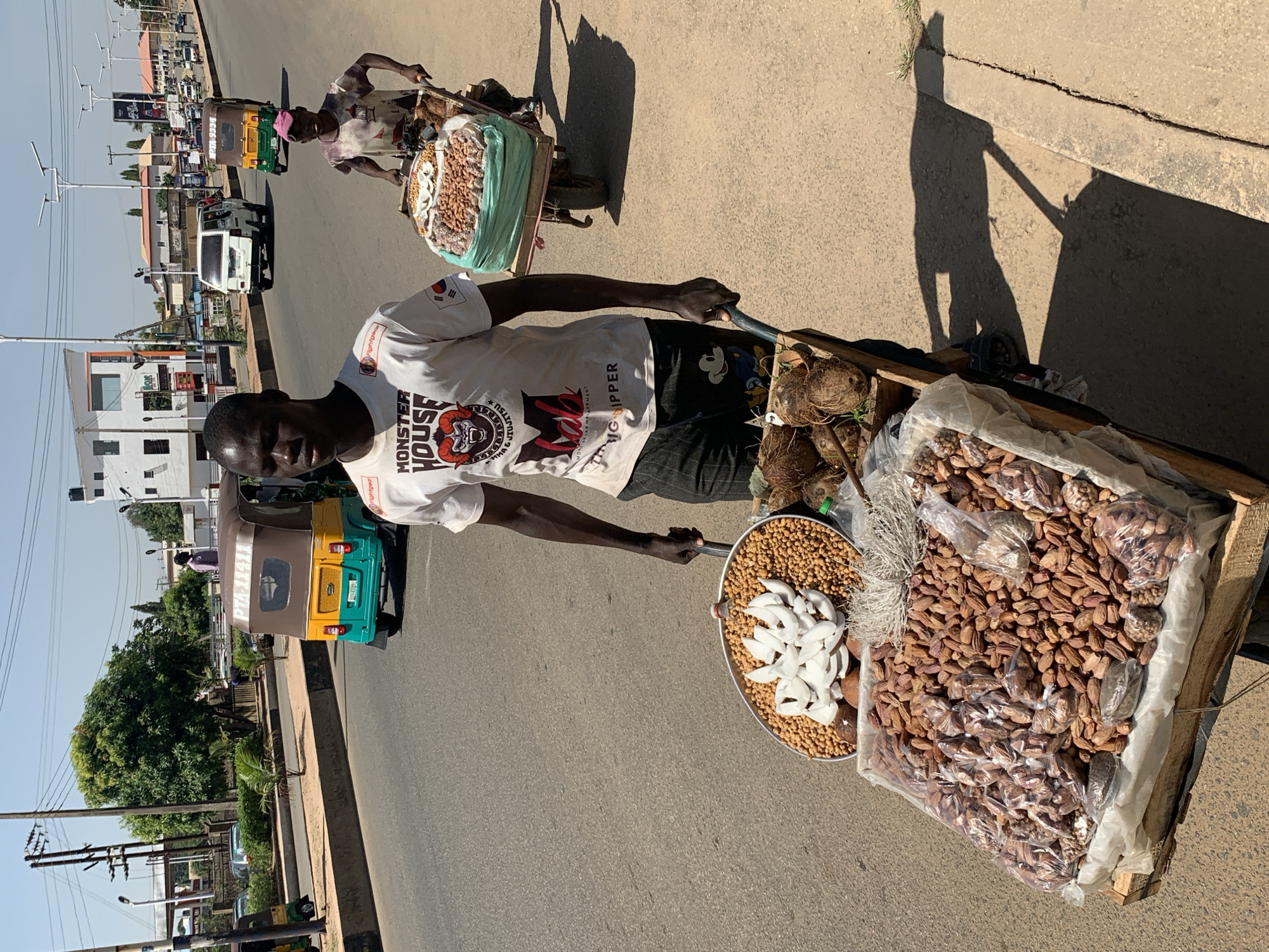 This image depicts a vibrant street scene in Gura Top, Nigeria, on a clear, sunny day. The strong, angled shadows suggest it is either late morning or early afternoon.
In the foreground, a young man, dressed in a white T-shirt and dark shorts, is pushing a wooden cart filled with goods for sale. His white T-shirt prominently displays the text "MONSTER HOUSE" and "KING DIPPER," along with a small patch resembling the South Korean flag on his left sleeve. The cart holds a diverse array of items: a large quantity of dates, many packaged in individual clear plastic bags, roasted peanuts or groundnuts displayed on a circular metal tray, and several pieces of peeled white coconut, also on the tray. A few whole coconuts are also visible near the front of his cart.
Further down the road, another street vendor, possibly an older woman wearing a patterned garment and a head covering, is also pushing a similar cart, laden with what appears to be more dates and nuts. The street is an asphalt road with faint markings, and various vehicles are present, including a white van and a distinctive green, yellow, and brown auto-rickshaw (known as a keke Napep in Nigeria). Along the roadside, there are several multi-story buildings, trees, and utility poles, indicating a busy urban or semi-urban environment. The overall scene captures daily commerce and street life.