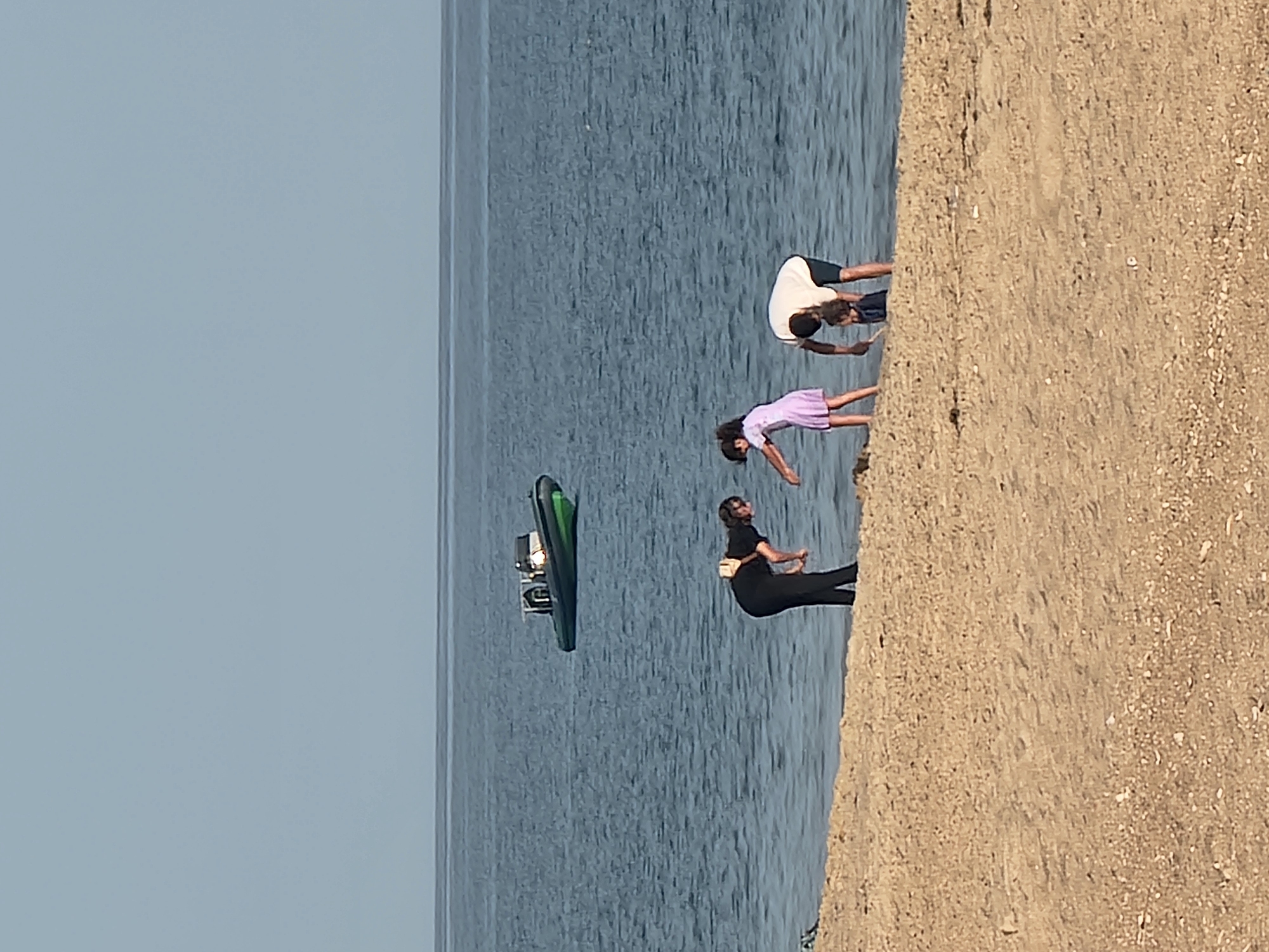 The image depicts a group of people by the sea on a sunny day, likely in Quarteira, Portugal. The scene is divided by a sandy embankment that slopes down towards the water. In the water, a dark green jet ski is visible with a small white motor.
On the embankment and at the water's edge, there are several individuals. Closest to the camera, a person in black activewear is seated with their legs bent. Further out in the water, three figures are partially submerged and appear to be engaged in a synchronized activity, possibly yoga or stretching, as they are all bending forward with their hands extended towards the water. One person wears a light purple dress, another a white t-shirt and dark shorts, and the third is mostly obscured. The water is a deep blue, indicating it is likely the ocean. The sky is clear and pale blue, suggesting good weather and a daytime setting. The sand is light brown and appears dry, with some small pebbles visible. There is no visible text in the image.
