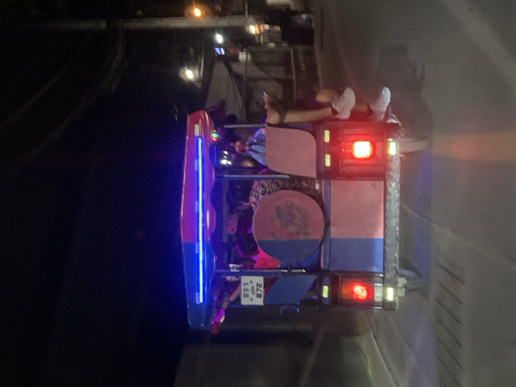 The image shows a colorfully lit tuk-tuk on a street at night. The vehicle is decorated with blue neon lights and red brake lights. People are riding in the back, with their feet visible towards the rear. The license plate is partially visible. The surrounding environment suggests an urban setting at night, though specific landmarks are not discernible.