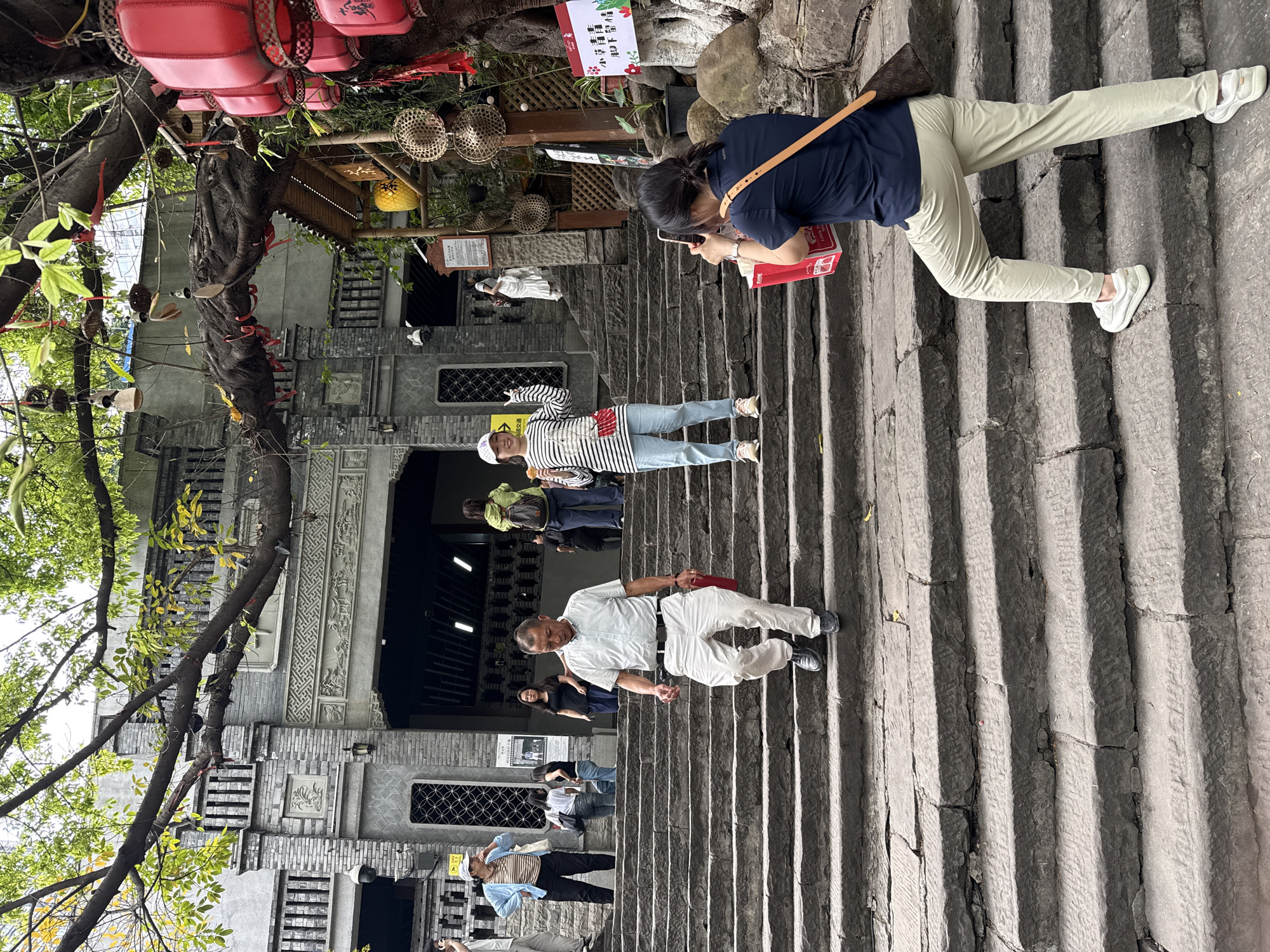 The image depicts a stone staircase in the city of Yuzhong District, China. Several people are walking up or down the stairs. A woman on the upper part of the staircase appears to be taking a photograph with her phone. The architecture includes stone buildings with ornate carvings, windows with geometric patterns, and hanging decorations such as lanterns and red ornaments attached to tree branches. The environment is partially shaded by trees.