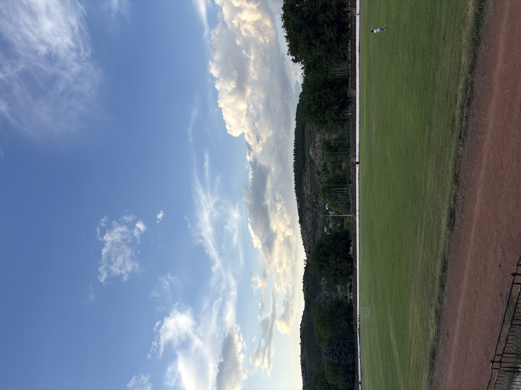 The image shows a wide shot of a green field, likely a sports field or stadium, in Boboshevo, Bulgaria. The field is bordered by a reddish-brown track in the foreground and a line of trees and hills in the background. The sky is blue with scattered white clouds. There are a few distant figures on the field, including one person in the lower right corner who appears to be walking or jogging. A soccer goal is visible in the distance on the left side of the field. The overall impression is one of a serene, open-air setting.