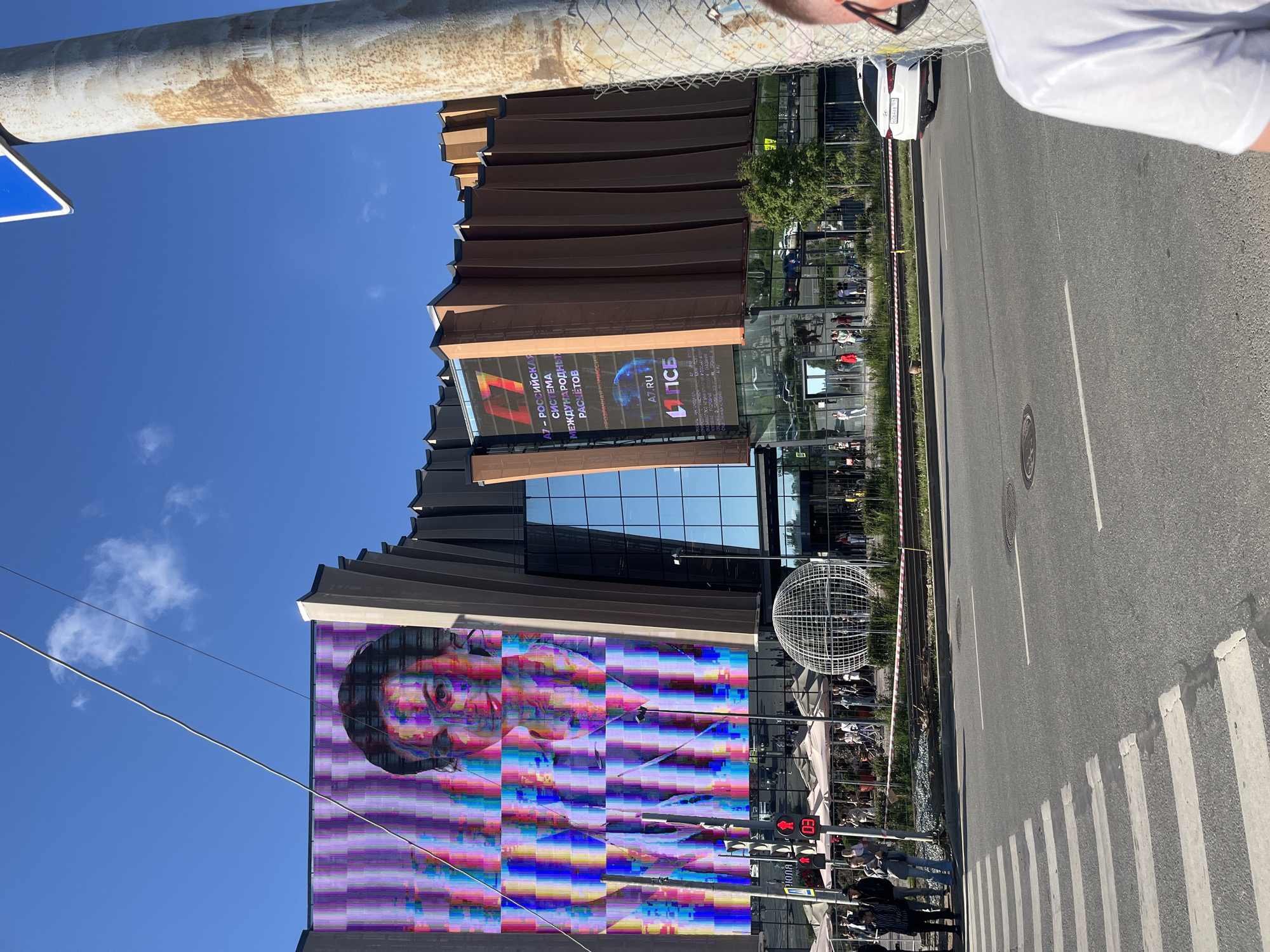 The image depicts a street scene in front of a modern, multi-story building with a large LED screen displaying a glitchy portrait. The building features a unique architectural design with a facade composed of angular, brown panels and large glass sections. To the left of the main screen, a smaller screen shows a logo and text that appears to be in Russian, possibly advertising a financial service.
In the foreground, a street with a pedestrian crossing is visible. Traffic lights show a red signal with the number '03' displayed on a digital counter below. Several people are seen walking on the sidewalk and near the building entrance. A large, spherical metal sculpture, resembling a globe, is positioned on the sidewalk.
The environment is a city setting under a clear blue sky with a few wispy clouds. A lamppost with a rusted surface and a blue sign is prominent in the right side of the frame, partially obscuring the building. The overall impression is a bright, sunny day in an urban area. The text on the screen and traffic light suggest the location is Russia, likely Perm, Russia, given the presence of Russian text and the typical urban infrastructure. A white sedan is parked on the right side of the road.