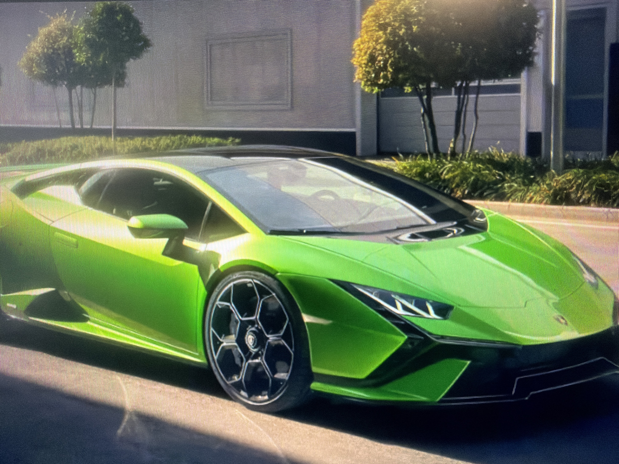 The primary subject of the image is a vibrant, lime green Lamborghini Huracan, likely an Evo model, positioned outdoors. The sports car is shown from a front-side angle, highlighting its aggressive styling, sharp lines, LED headlights, and distinctive hexagonal-spoked wheels with a subtle Lamborghini logo on the central hub.
The car is stationary, parked on what appears to be a paved surface, possibly a road or parking lot. The setting suggests an urban or suburban environment, characterized by a modern building in the background. This building features a light-colored, textured wall with a prominent rectangular architectural detail resembling a framed recess or window. Several well-maintained trees with dense, rounded canopies and lush green shrubs line the base of the building, indicating a landscaped area.
The time of day is clearly daytime, with strong, natural sunlight illuminating the scene. This bright light creates prominent highlights on the car's reflective surface and casts distinct shadows on the ground, suggesting sunny and clear weather conditions. There is no specific activity or event taking place, beyond the presentation of the car. No visible text is discernible in the image apart from the car's brand logo on the wheel. The overall impression is one of luxury, modernity, and high performance.
