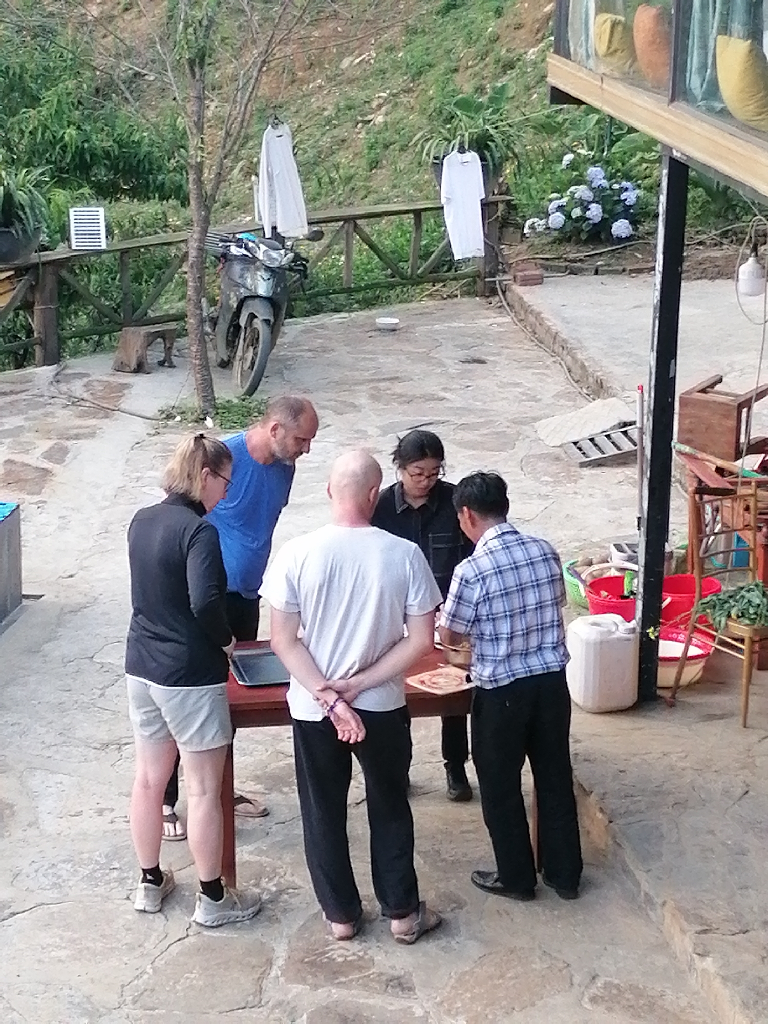 Five individuals are gathered around a wooden table in an outdoor, rustic setting, likely a guesthouse or home in Sa Pa Ward, Vietnam. The scene is illuminated by natural daylight, suggesting late morning or afternoon.
On the left, a woman wearing a black jacket, light shorts, and sneakers stands with her back to the viewer, looking down at the table. Next to her, a man in a blue t-shirt observes the table. In the center, another man in a white t-shirt and dark pants stands with his hands clasped behind his back, also looking at the table. To his right, a woman in a dark shirt and glasses peers down, while a man in a blue and white plaid shirt appears to be actively involved with something on the table, possibly stirring or preparing food in a metal tray or pan. Their collective focus indicates an activity or demonstration taking place on the table.
The ground is a mix of irregular stone paving and concrete. In the background, lush green hills and trees rise. A black motorcycle is parked near a slender tree, with two white t-shirts hanging to dry on a wooden railing nearby. Another white t-shirt hangs on a stand further right. A white ventilation-like object sits on the railing. Adjacent to the gathering, there's a building with glass windows, displaying colorful cushions inside. Near the building, a cluster of light blue hydrangeas adds a touch of color, and various household items like red buckets, a white plastic container, and wooden furniture are visible. There is no discernible text in the image.