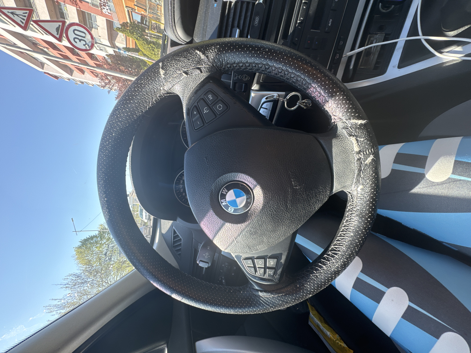 The image captures a view from inside a car, focusing primarily on the steering wheel, which features a BMW logo at its center. The steering wheel appears worn and slightly damaged, particularly around the rim where the leather or material is peeling off.
Beyond the steering wheel, the dashboard and interior components of the vehicle are visible, suggesting a car with a navigation system and control buttons. An iPhone charging cable hangs below the dashboard.
Outside the car, through the windshield, there's a glimpse of an urban environment. Several road signs and a building are visible, indicating a street in a town or city. Specifically, based on the road signs (pedestrian crossing, speed limit 30km/h, hump), this image was likely taken in the city of Sofia, Bulgaria.