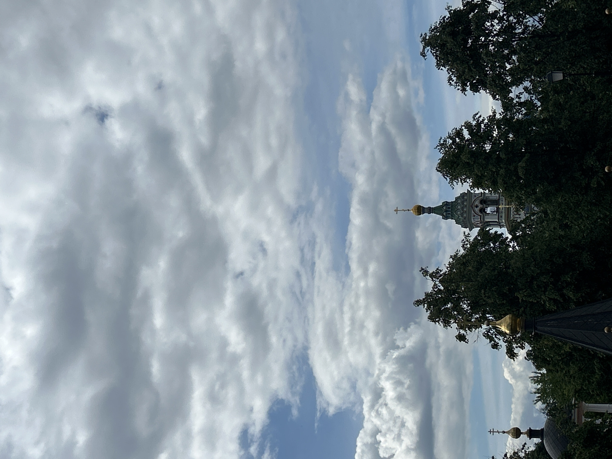 The image shows a partial view of a church or religious building amidst trees, against a cloudy sky. The architectural details are intricate, with golden domes topped with crosses. The location is the city of Unknown, Unknown.