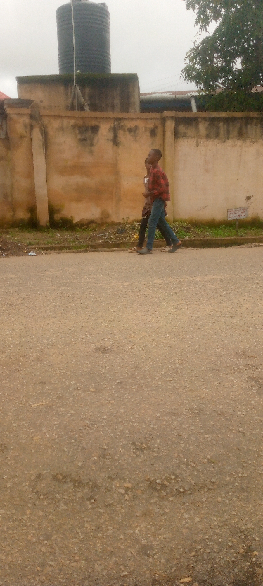 Two young males walk side by side on an asphalt road. The male in the foreground is wearing a red and black plaid shirt and blue jeans, while the male behind him is wearing a brown shirt and dark pants. They are walking past a beige concrete wall with some moss and dirt on it. Behind the wall, a large black water tank is visible, along with some trees and buildings. The sky is overcast and the overall lighting suggests a daytime scene. The road surface is textured with small stones and appears to be a typical urban or semi-urban environment.