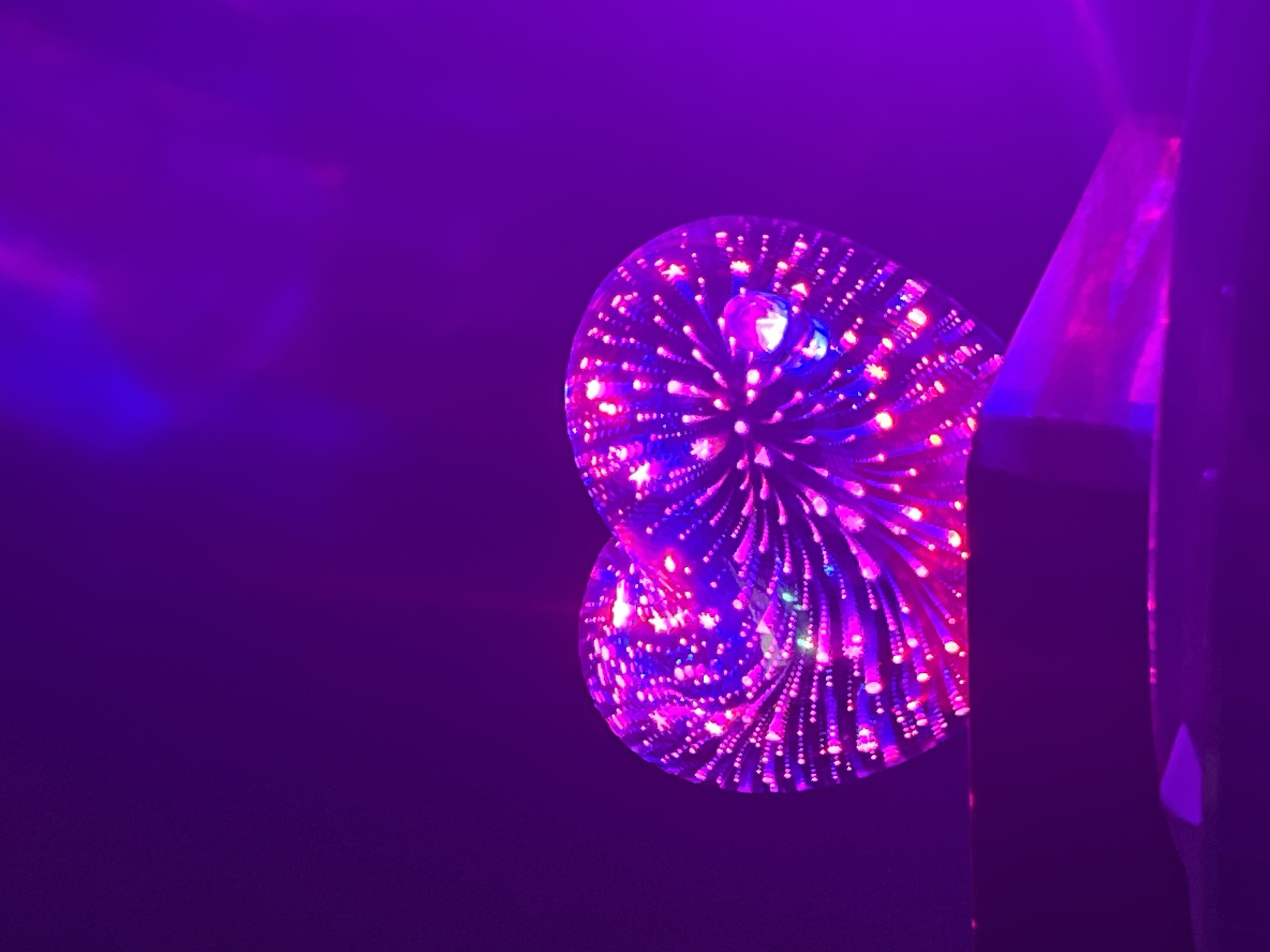 A close-up, low-angle shot shows a heart-shaped object with internal pink and blue lights against a dark purple background. The object is positioned on a dark, flat surface, possibly a shelf or table, with a sliver of another object beneath it. The heart appears to be made of glass or a similar transparent material, with an intricate pattern of tiny lights embedded within. The lighting is dim and moody, creating a bokeh effect with the scattered lights. The overall impression is one of abstract beauty and artistic lighting.