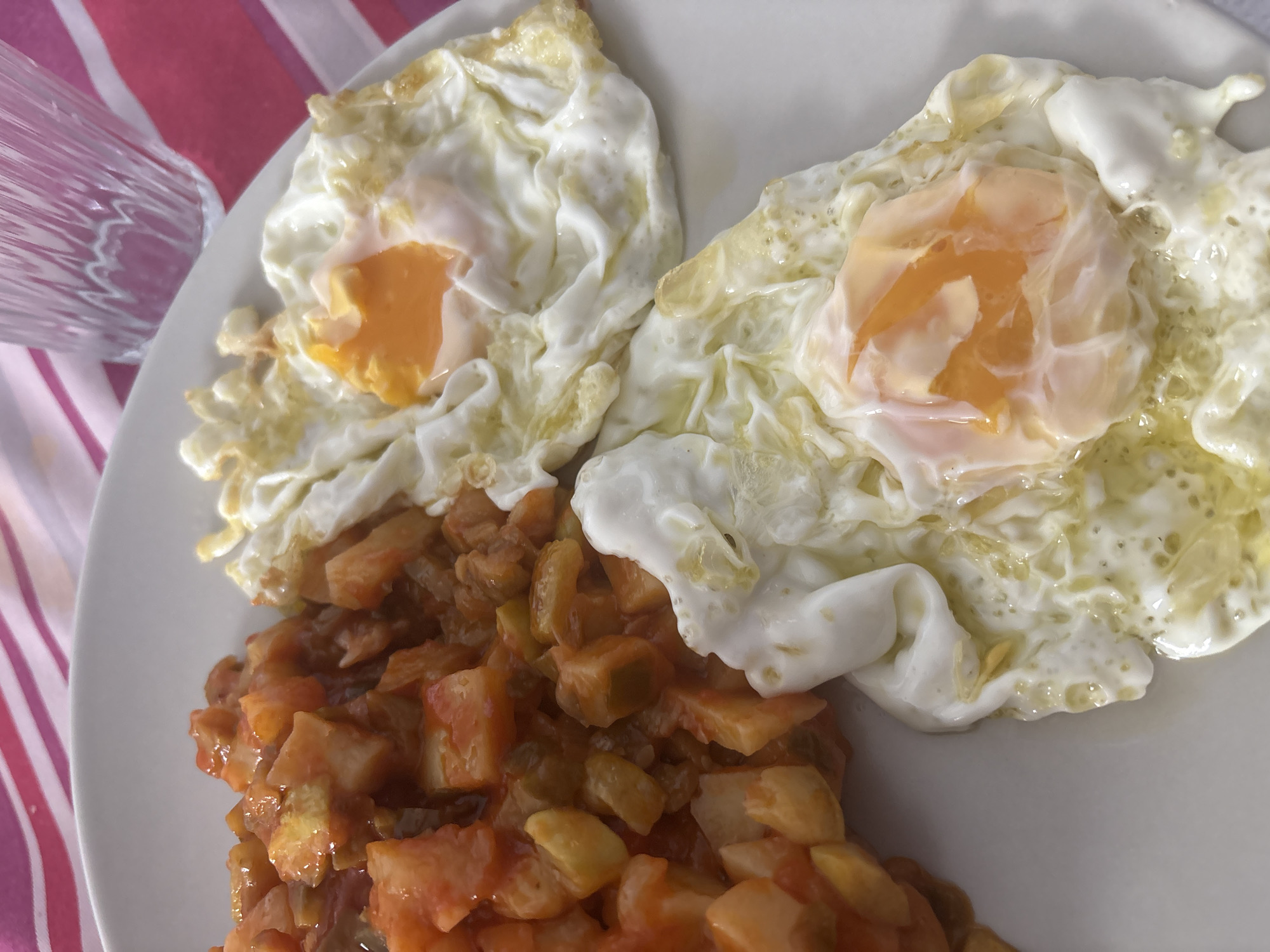 A high-angle, close-up shot captures a white ceramic plate holding a prepared meal, situated on a red and white striped tablecloth, indicative of a domestic setting within Urbanización Encinares De Alcolea, Spain. The plate features two sunny-side-up fried eggs positioned on the right, each displaying a vibrant orange, liquid yolk surrounded by firm, opaque white albumen with slightly crisp, browned edges. To the left, a heap of diced, reddish-orange and greenish-yellow ingredients, primarily cubed potatoes and possibly bell peppers and tomatoes, are coated in a dense, reddish-brown sauce. A clear, fluted glass containing a transparent liquid is partially visible in the upper right background, resting on the striped surface.