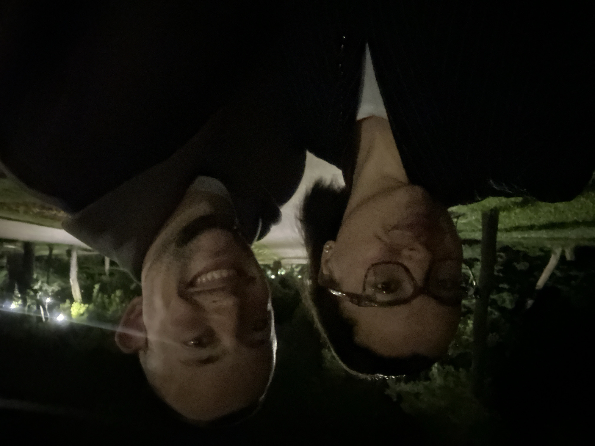 A low-light, wide-angle selfie captures a man and a woman posing outdoors at night, possibly in the Sea Garden in Varna, Bulgaria. The woman, on the left, wears glasses and a dark jacket over a white top. The man, on the right, has a shaved head and is smiling broadly. The background is dark, with some out-of-focus greenery and string lights visible. The image appears to be taken in a public park area with trees lining a pathway.
