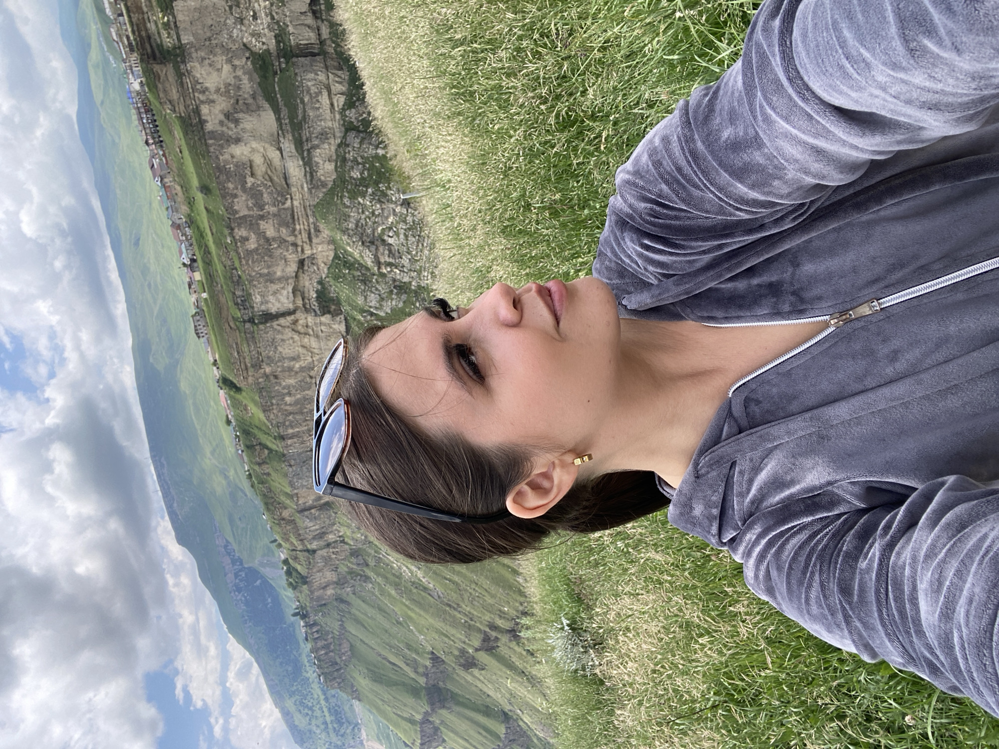 A selfie shows a young woman in a gray velour hoodie, with sunglasses perched on her head, outdoors in a grassy, mountainous area. The woman is facing to her right, with her left shoulder and part of her chest visible. Her hair is dark brown and pulled back into a ponytail. She is wearing a small gold earring in her left ear. The background features a dramatic mountain landscape with steep cliffs, green slopes, and a village nestled on the cliffside. The sky is partly cloudy. The location is identified as the village of Арани, Russia.