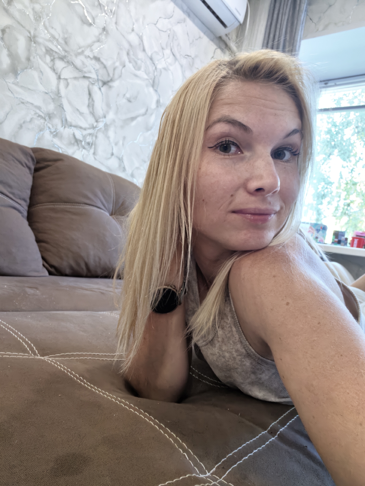 A blonde woman with fair skin and freckles is lying on a brown couch, looking towards the camera. She has blonde hair, and her makeup includes eyeliner and mascara. She is wearing a grey tank top. Her left arm is bent, with her hand resting on her chin. A black smartwatch is visible on her left wrist. The couch is upholstered in brown fabric with white decorative stitching. In the background, a wall with a marble-like texture is visible, along with a white air conditioner unit. A window with trees outside is also visible. The scene appears to be indoors, possibly in a residential setting in the city of Sterlitamak, Russia.