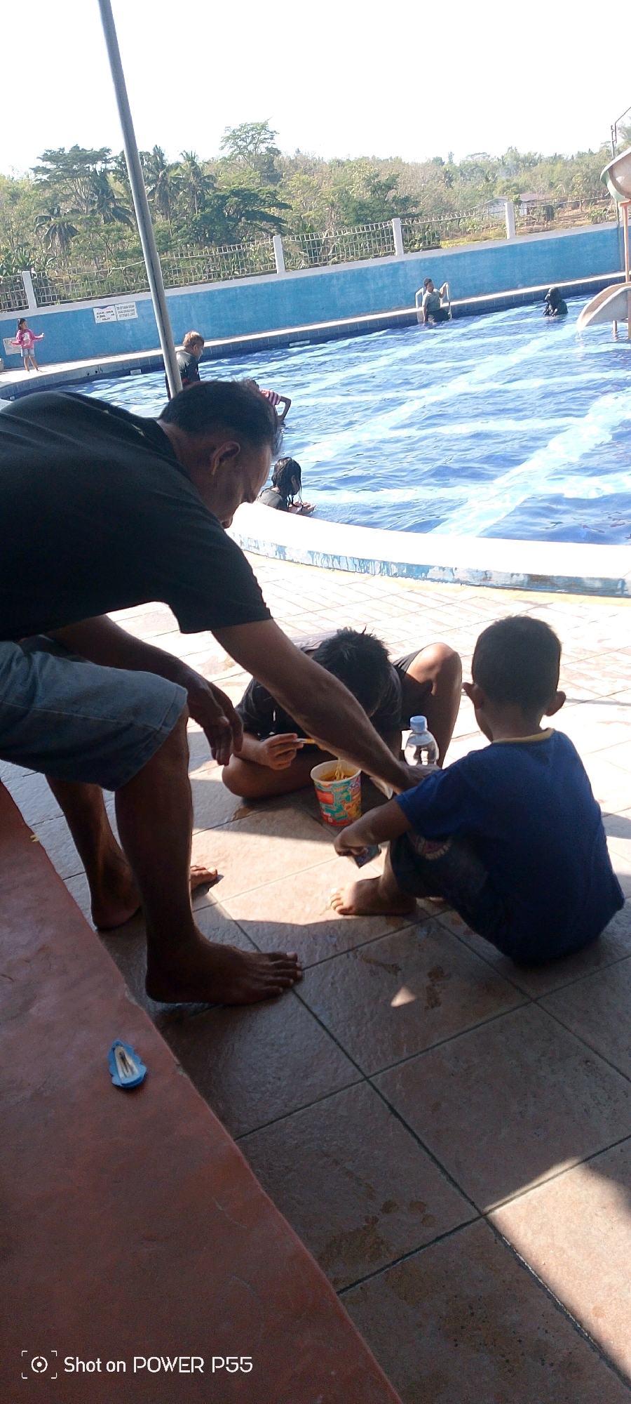 The image shows a group of people by a swimming pool on a sunny day. In the foreground, a man wearing a black t-shirt and denim shorts sits on a red bench, leaning towards two children. He is offering food to one of the children, who is wearing a blue t-shirt. The other child, wearing a black t-shirt, is also near the man. There is a bowl of noodles and a bottle of water on the tiled ground. In the background, the swimming pool is filled with blue water and several people are swimming or standing in the pool. A white fence surrounds the pool area, and trees and foliage are visible beyond the fence. The scene appears to be in an outdoor recreational area.