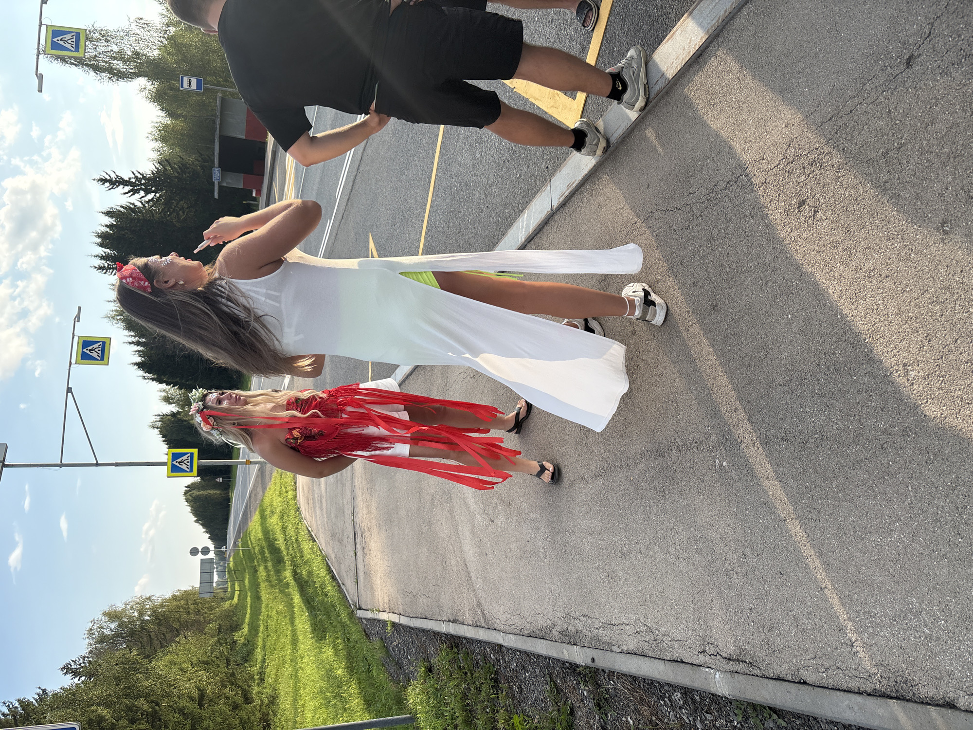 Three individuals are standing on a paved pedestrian crossing in Goldyrevskii, Russia. The person on the left wears a red, white, and floral outfit, adorned with a flower crown and red ribbons. The middle person is dressed in a long white dress with a high slit revealing neon green shorts underneath, and a red bandana on their head, holding a lip gloss to their mouth. The person on the right is wearing black shorts and a black t-shirt. A bus stop shelter is visible in the background, along with a road and a line of trees. The sky is blue with scattered clouds, and the sun is casting long shadows.