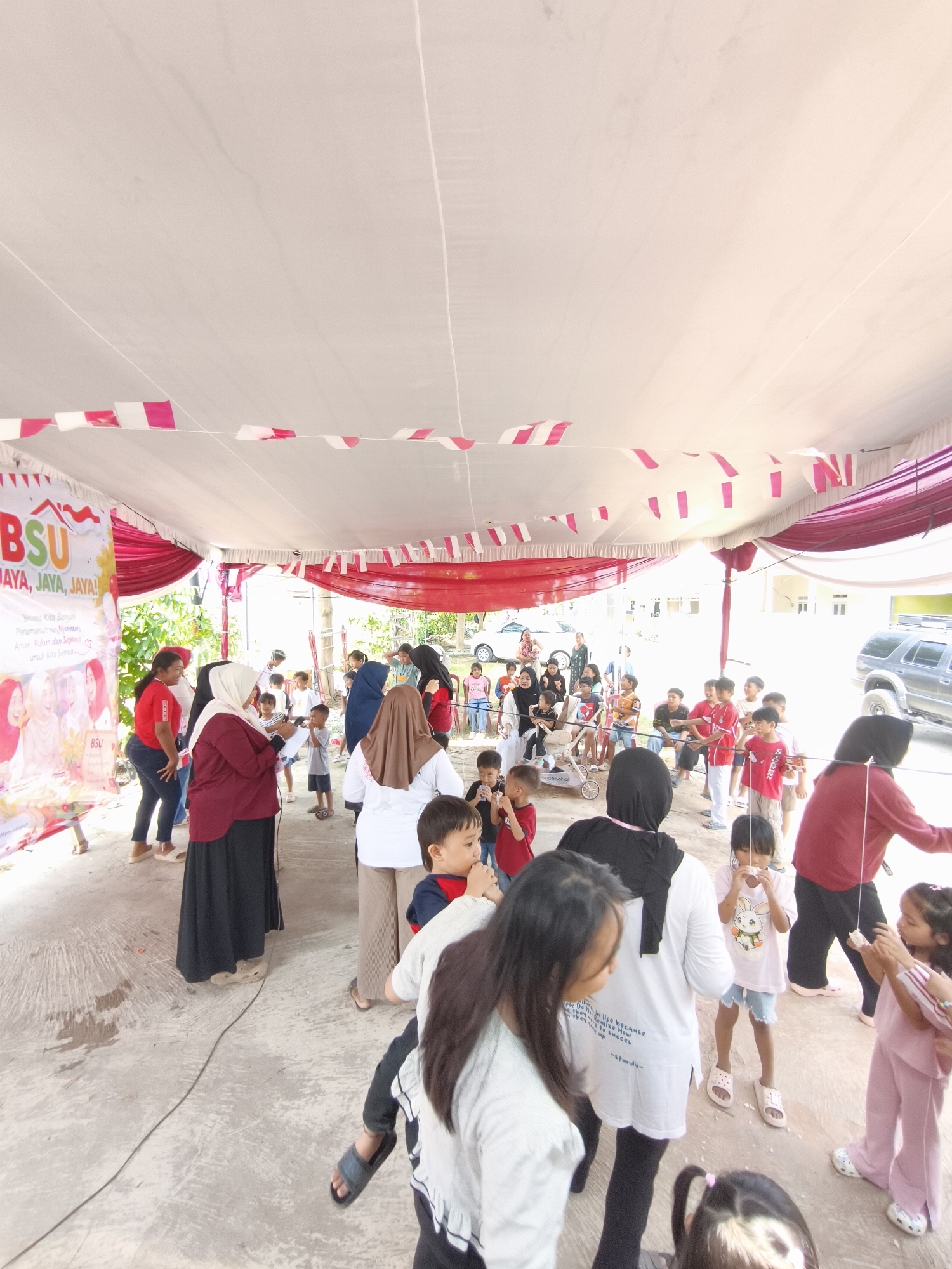 The image depicts an outdoor gathering under a white tent decorated with red and white streamers. A banner for "BSU" with the slogan "JAYA, JAYA, JAYA!" is visible on the left. A crowd of people, predominantly children and adults, are participating in a game or activity. Many children are holding strings with objects attached, and some appear to be engaged in a competition. Adults are supervising and interacting with the children. The ground is paved concrete, and in the background, there are trees, buildings, and parked vehicles, suggesting an urban or semi-urban setting within Bandar Lampung, Indonesia. The overall atmosphere appears festive and communal.